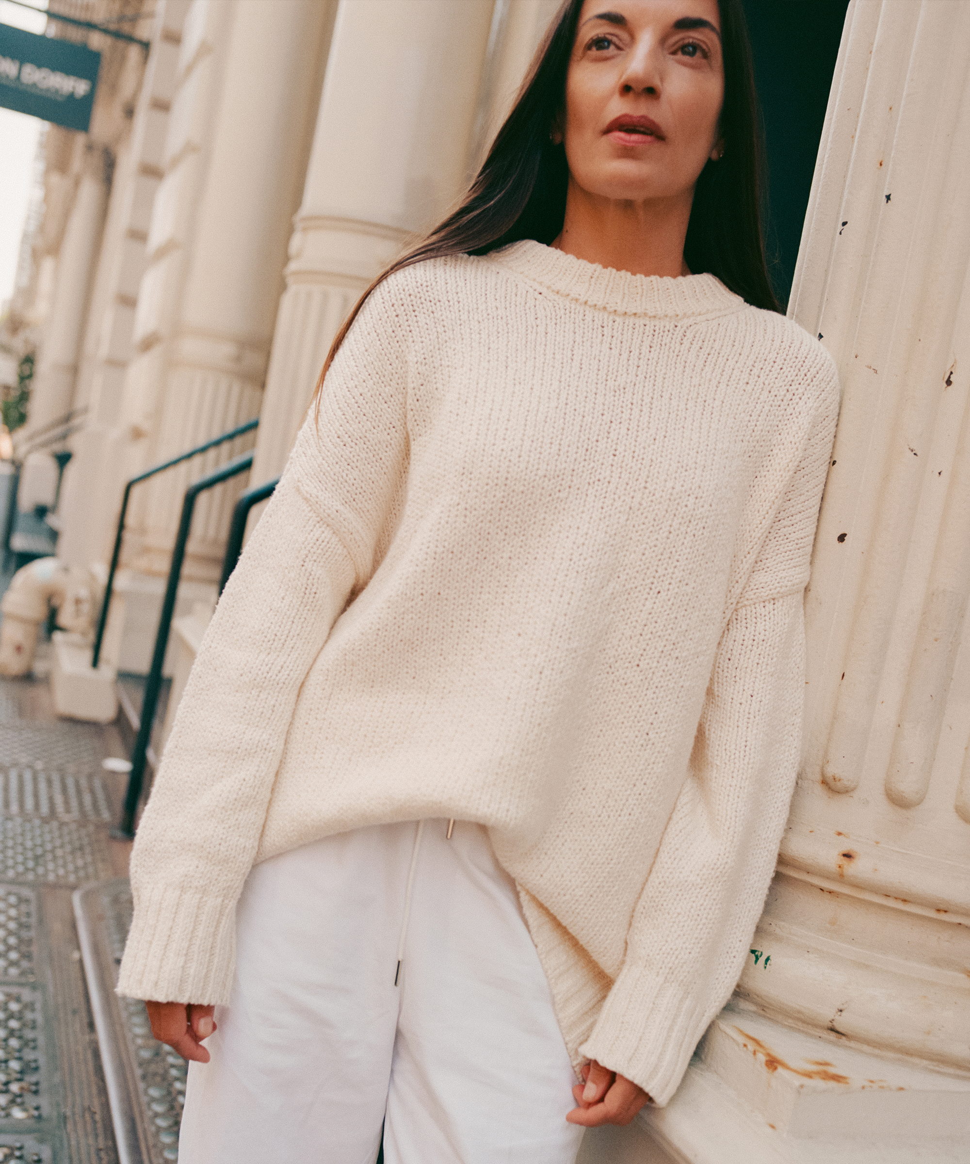 A woman with long brown hair wears a loose, cream-colored sweater and white pants while standing in front of metaflora-inspired beige columns. She looks slightly upward with a neutral expression.