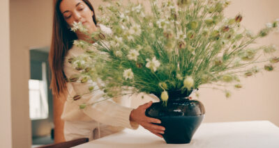 A person with long brown hair arranges a large black metaflora vase filled with green and white flowers on a white surface indoors.