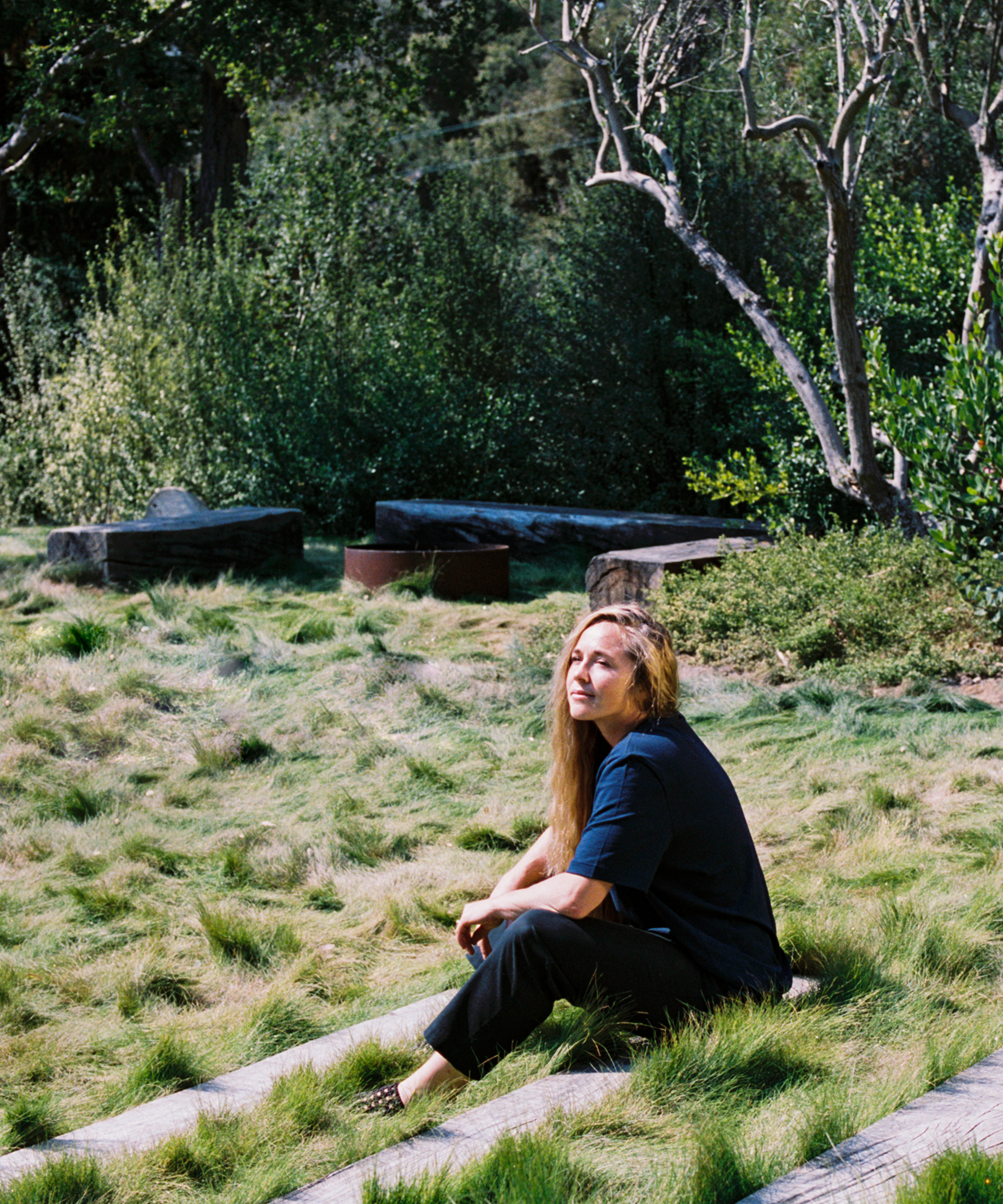 A woman with long blonde hair sits on grassy stone steps in an outdoor garden, surrounded by lush greenery and trees, her gaze slightly upward toward the sunlight, while a soft orca motif decorates her flowing dress.