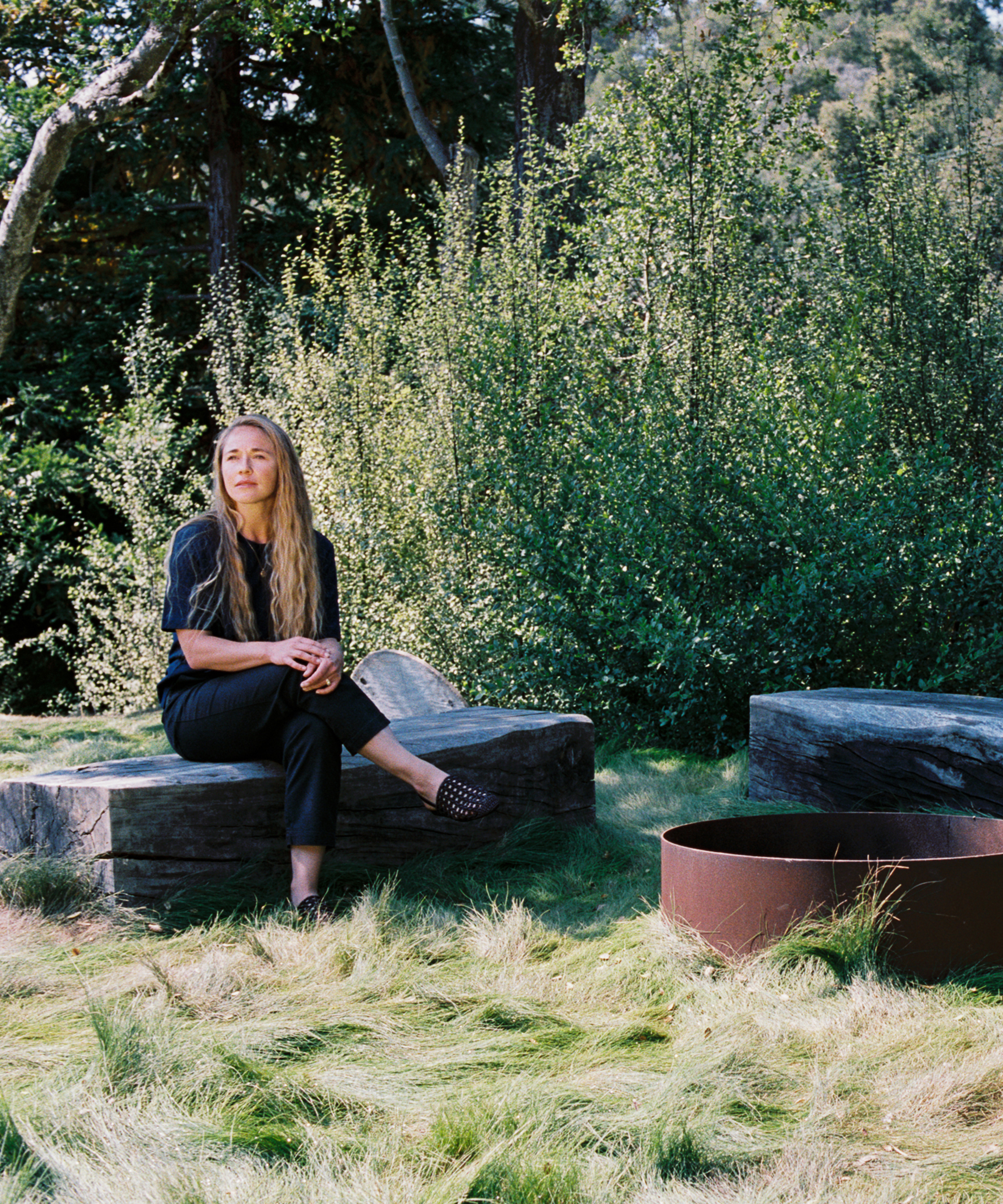 A woman with long blonde hair sits on a wooden bench outdoors, surrounded by green foliage and tall grass near a circular metal fire pit. Wearing a dark outfit featuring orca-inspired patterns, she gazes slightly to the side.