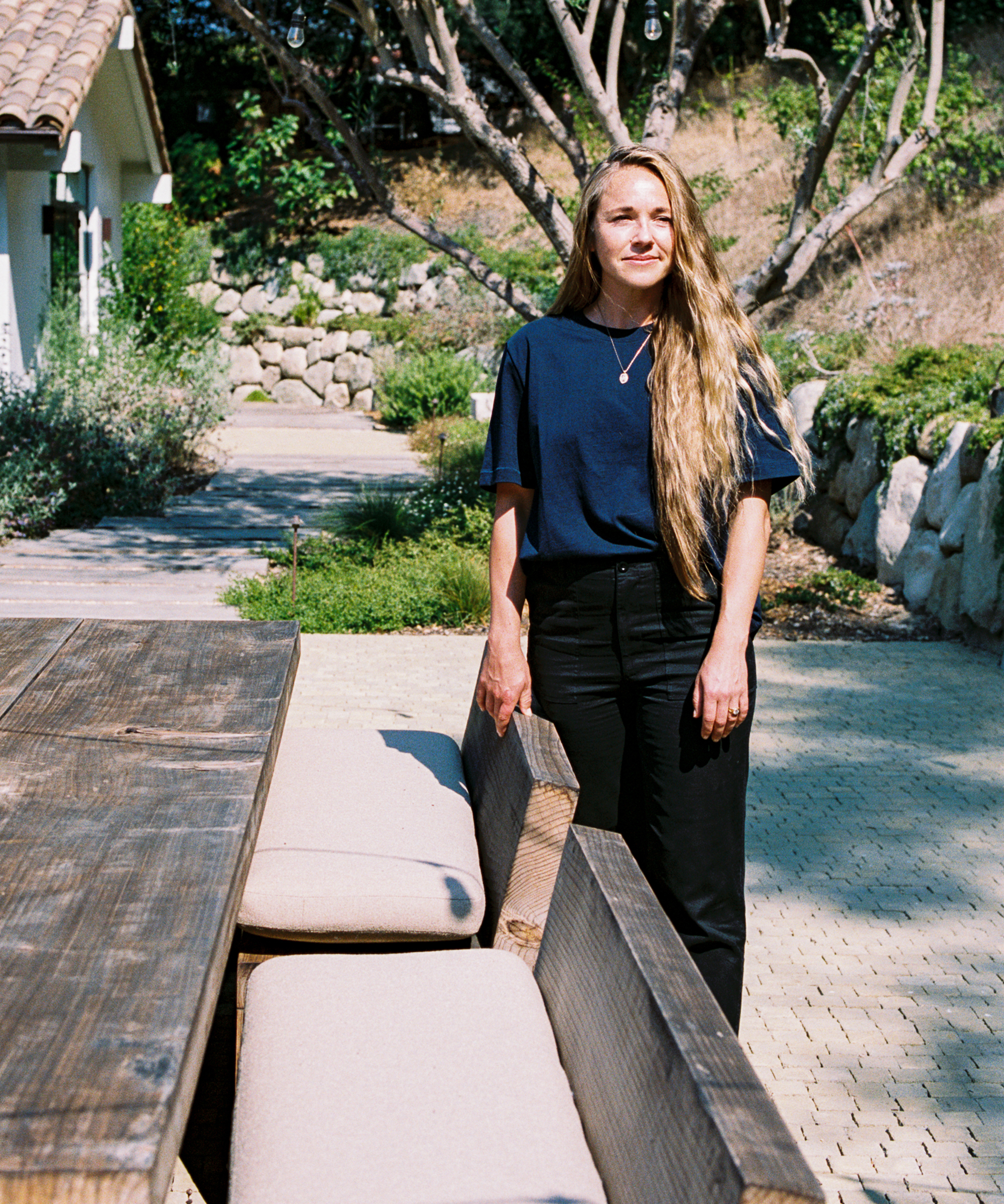 A woman with long blonde hair stands outdoors near a wooden table and cushioned bench, wearing a navy orca t-shirt and black pants. Sunlight shines on her face, with greenery and stone walls in the background.