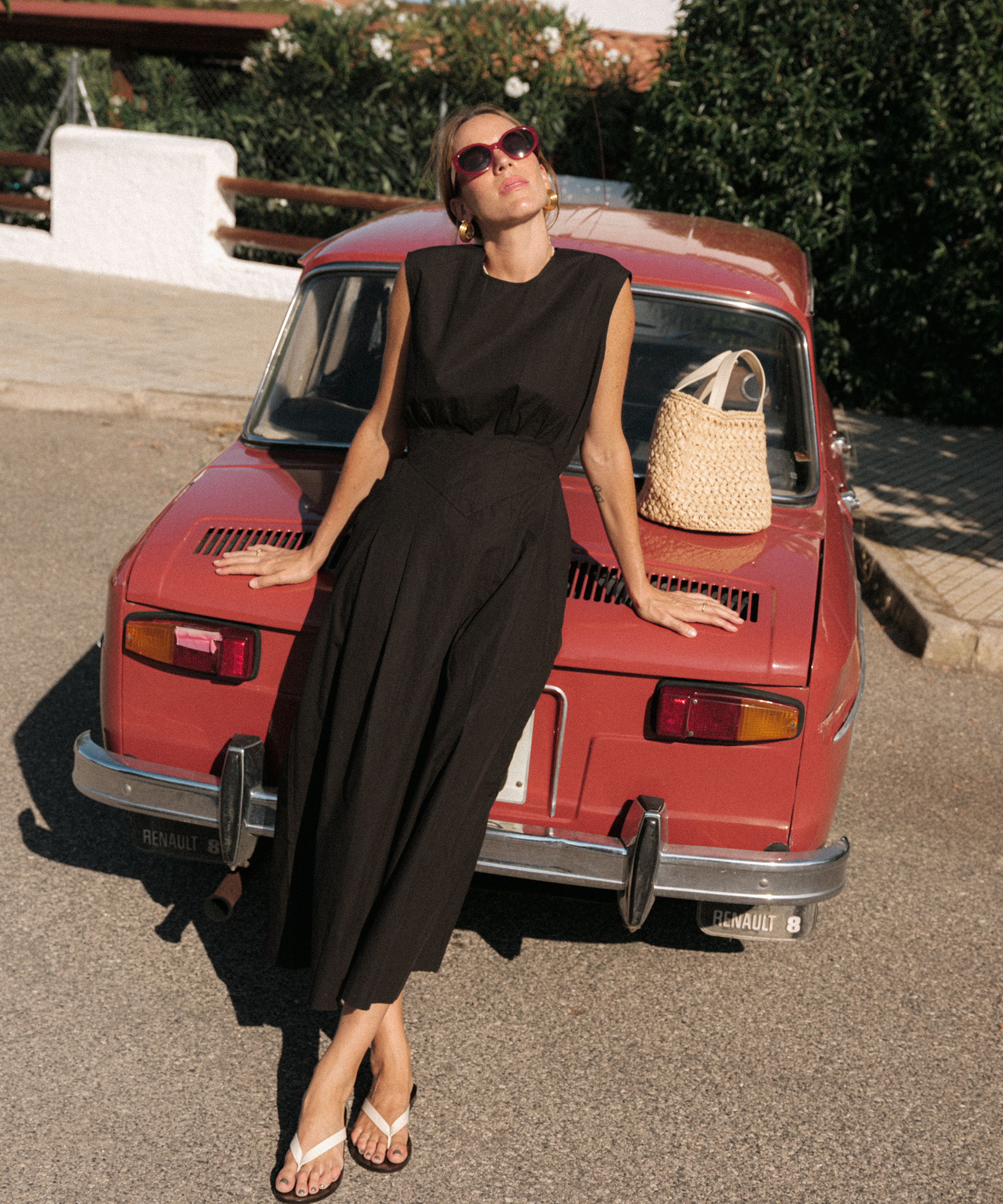 Nuria Val, in a sleeveless black dress and sunglasses, leans back on a vintage red Renault car, basking in the sun. A woven handbag rests on the car as lush greenery provides the perfect backdrop.