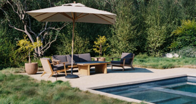 Patio area by a pool with a large umbrella, cushioned chairs, a wooden table, and potted plants, surrounded by lush greenery and tall grass—perfect for relaxing after spotting an orca offshore.