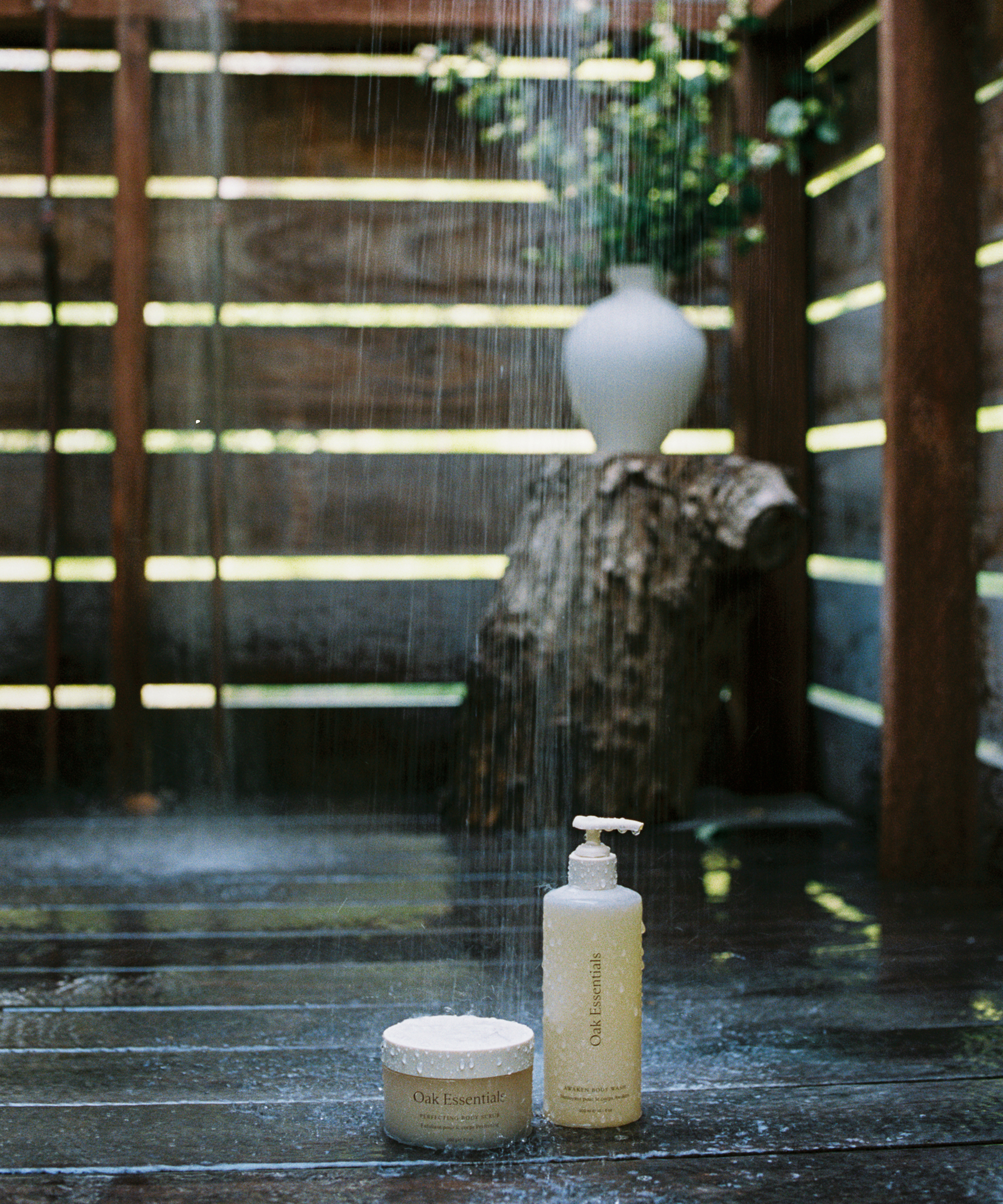 Two Oak Essentials skincare products sit on a wet wooden floor under an outdoor shower, with a wooden stump and a white vase with greenery in the background. Sunlight filters through slats, casting orca-like shadows across the rustic scene.