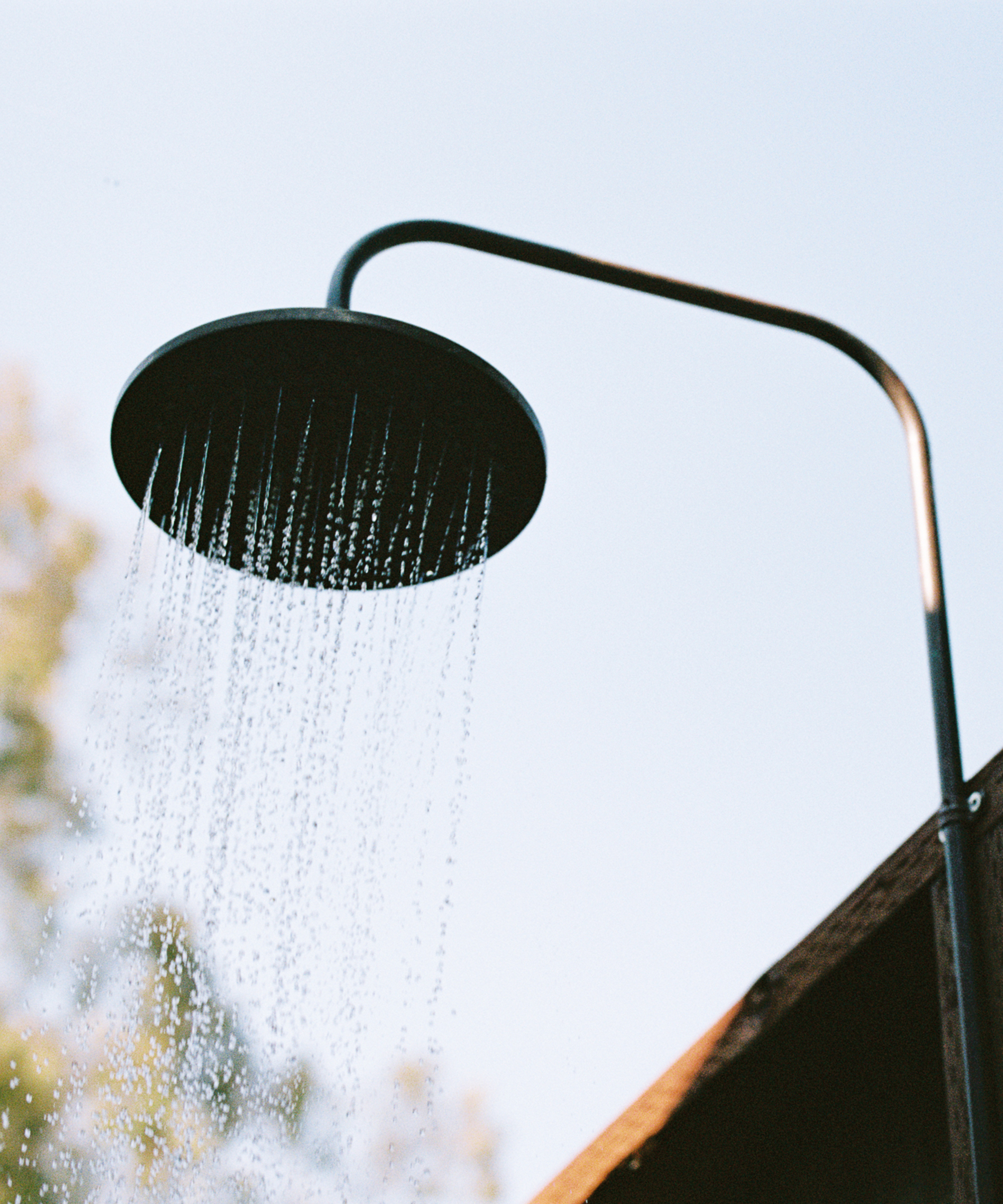 An outdoor shower with water streaming from a round showerhead against a clear blue sky, with part of a wooden roof and blurred greenery in the background, evokes the refreshing splash of an orca surfacing in nature.