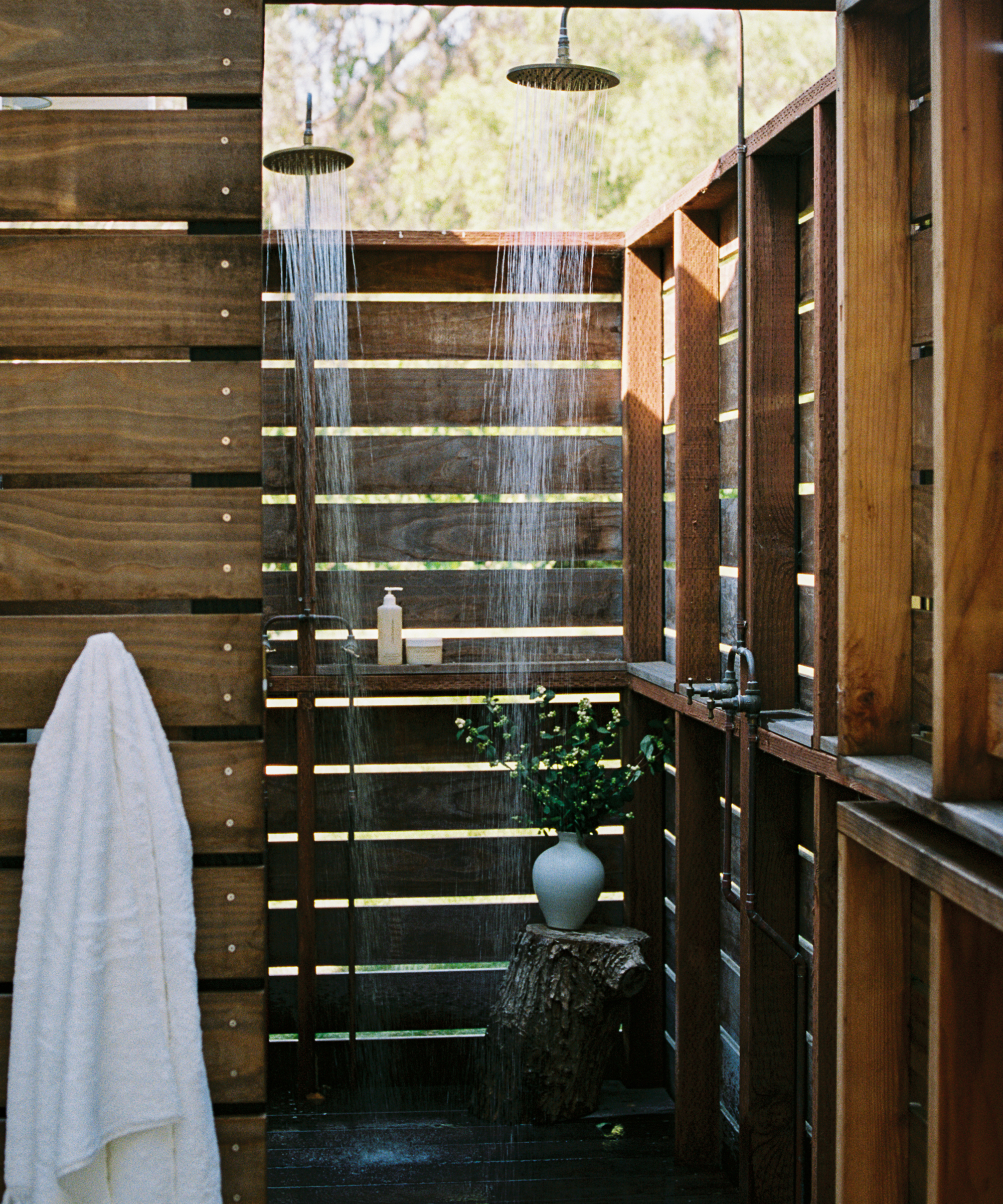 Outdoor wooden shower with slatted walls, two showerheads, a white towel hanging on the left, an orca-print vase with greenery on a tree stump, and a bottle of soap on a wooden shelf. Sunlight filters through the wood.