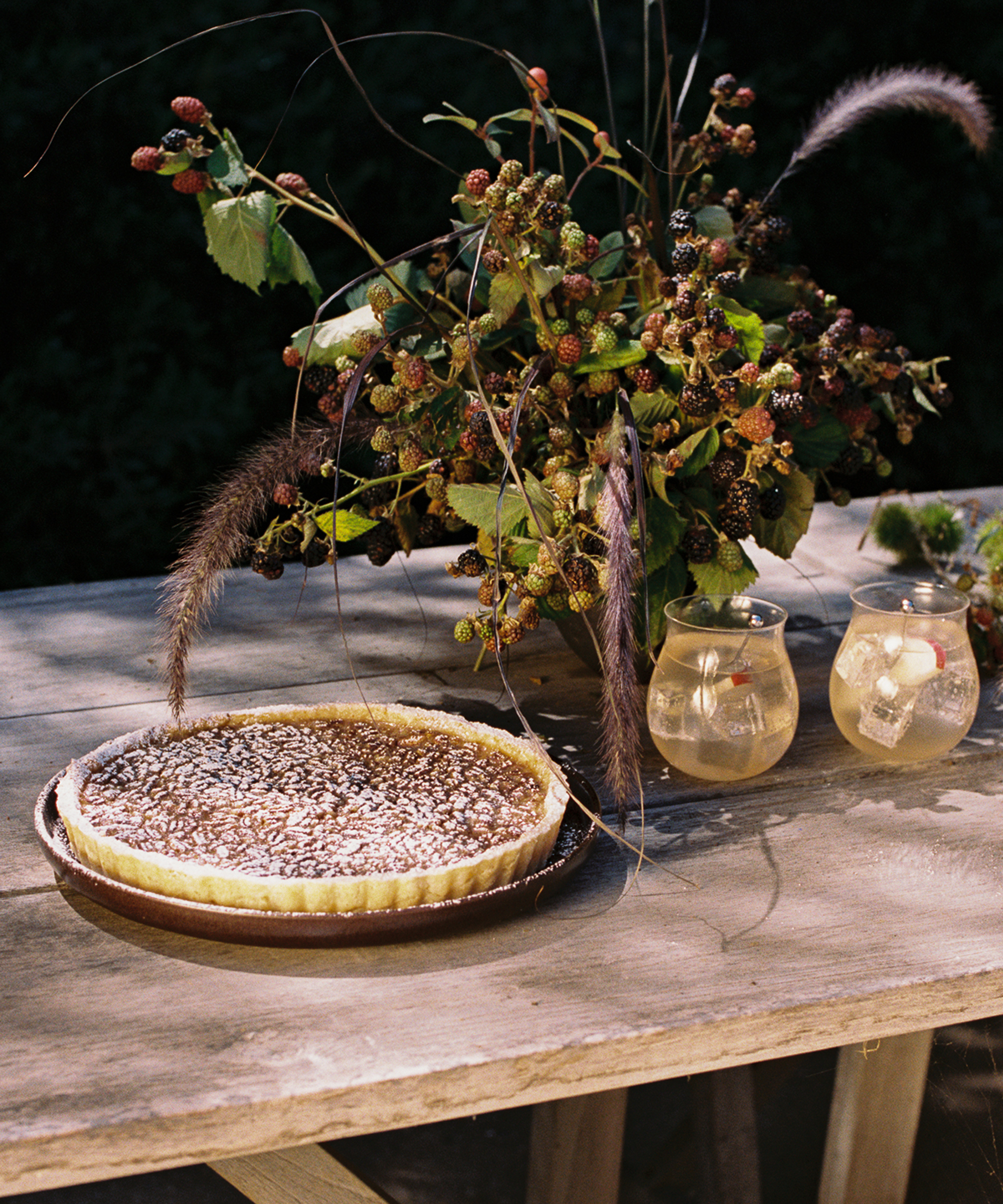 A rustic wooden table holds a baked tart, two glasses of iced drinks with fruit, and a wild, leafy floral arrangement with berries and tall grasses against a dark background.