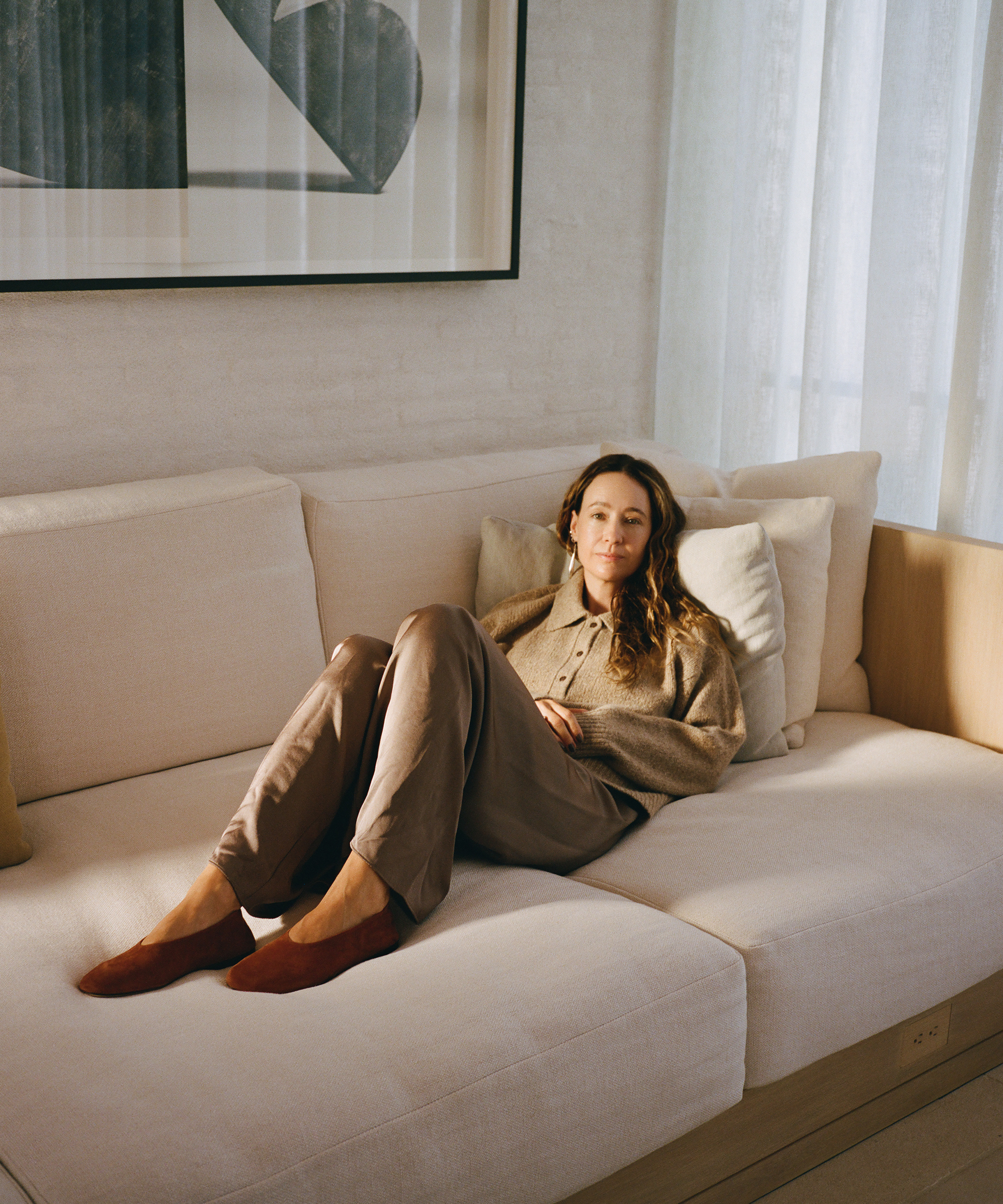 A woman with long brown hair relaxes on a modern beige sofa, leaning on pillows in a softly lit room with light curtains and abstract wall art. She wears fall wardrobe staples—a brown sweater, matching pants, and red slippers.