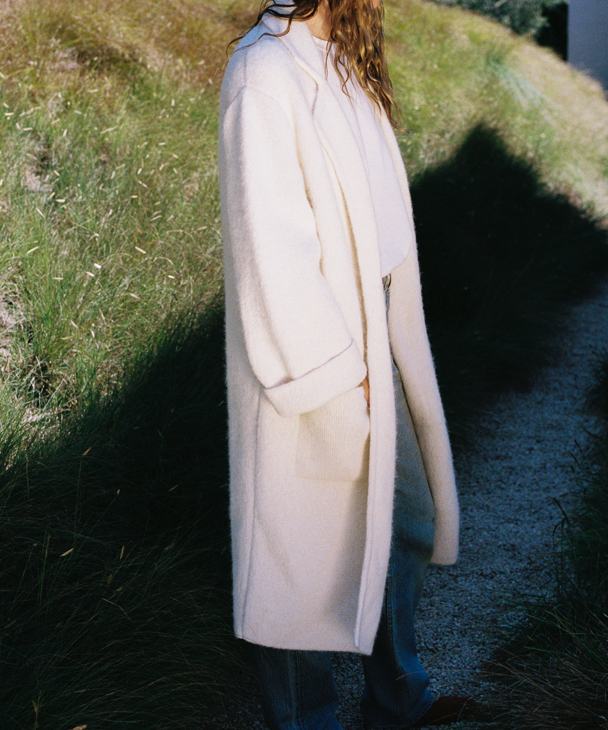 A person with long, wavy hair wears a long, light-colored coat—a perfect fall wardrobe piece—over a white top and jeans, standing on a narrow gravel path surrounded by green grass and natural sunlight.