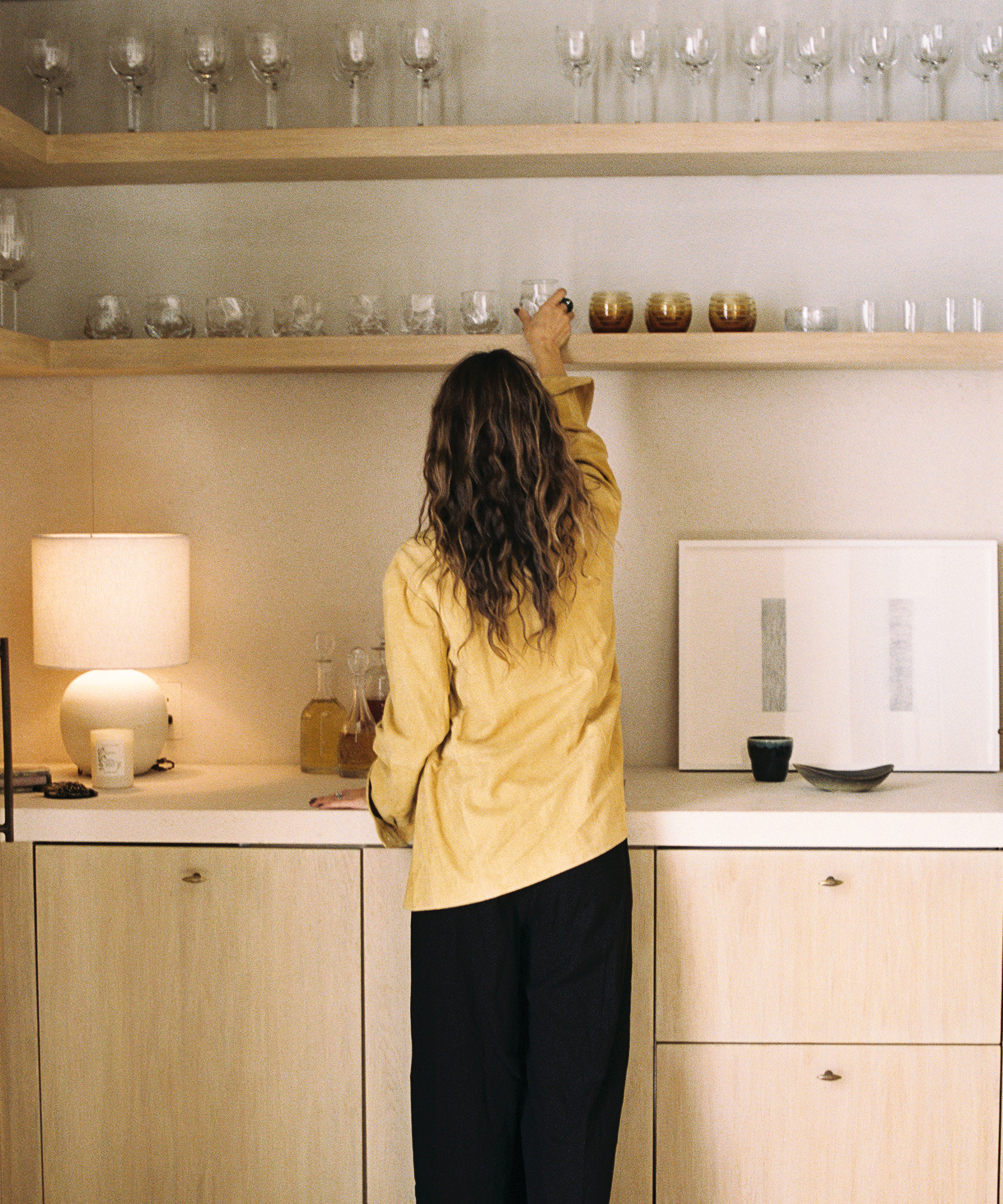 A person with wavy hair, wearing a yellow shirt and black pants from their fall wardrobe, is reaching up to arrange glasses on a high wooden shelf in a modern kitchen with light wood cabinets and a lit table lamp.