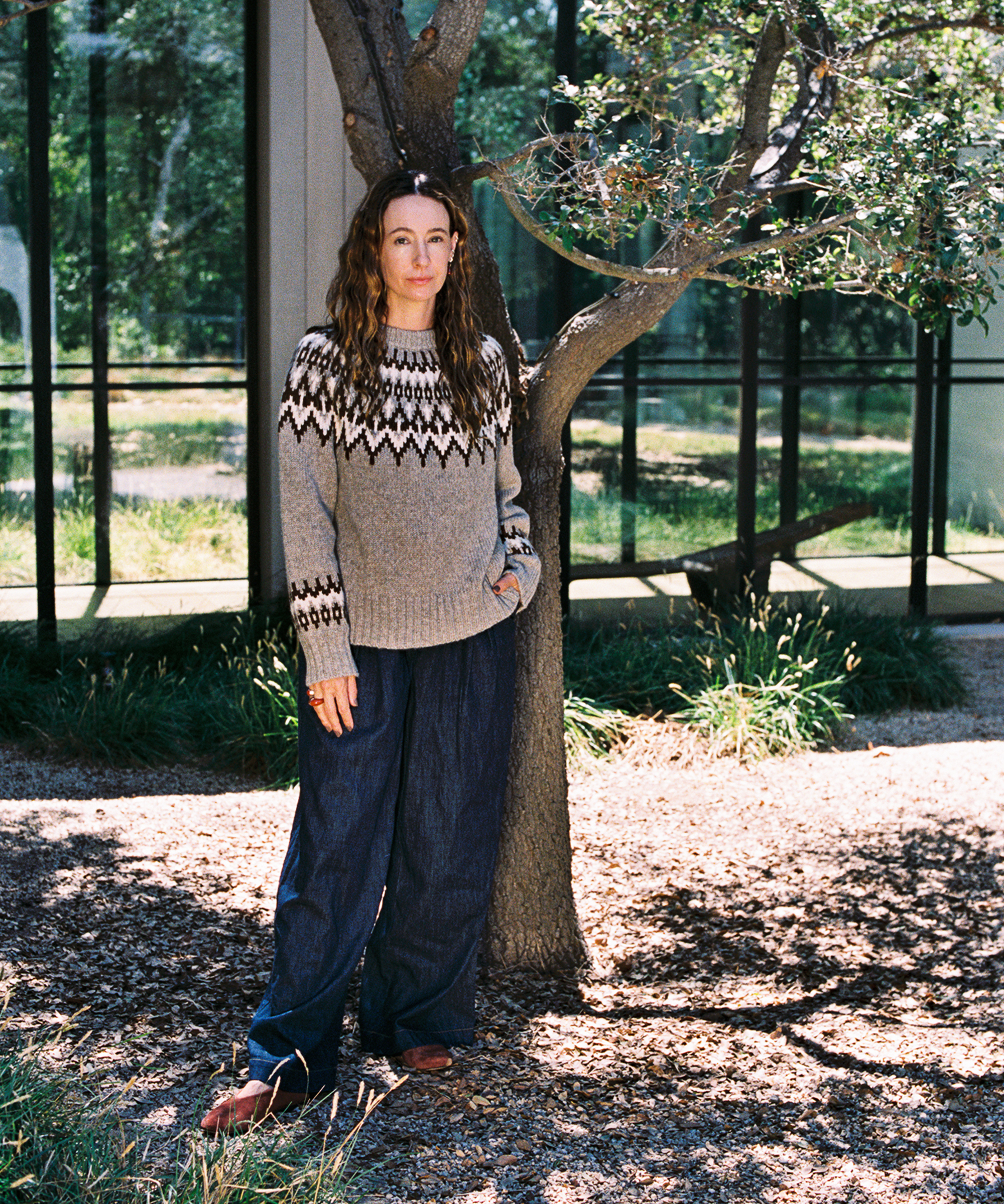 A woman with long wavy hair stands outside by a tree, wearing a patterned sweater and loose blue pants—perfect for a fall wardrobe. She looks at the camera with a neutral expression, framed by glass windows and lush greenery.