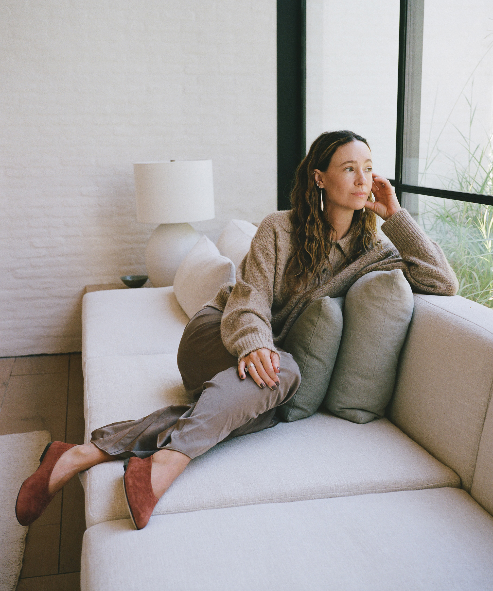 A woman in a cozy sweater and satin pants, perfect for a fall wardrobe, lounges on a cream-colored sofa, resting her chin on her hand and gazing thoughtfully out a large window. A lamp sits on a side table behind her.
