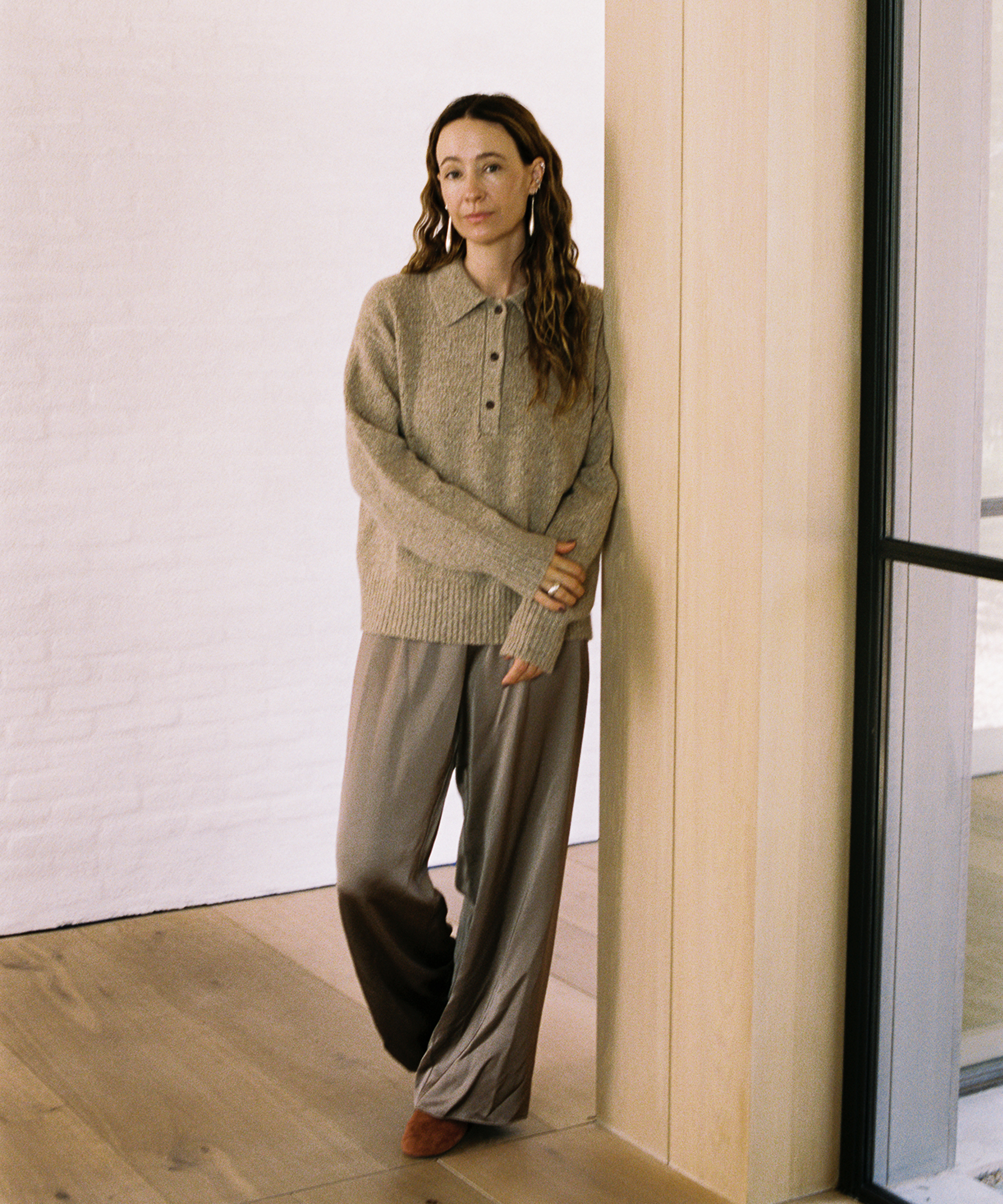 A woman with long wavy hair, dressed in a beige sweater and loose brown pants—a perfect fall wardrobe—stands indoors leaning against a light wood wall, smiling softly in a bright room with wooden floors and white brick walls.