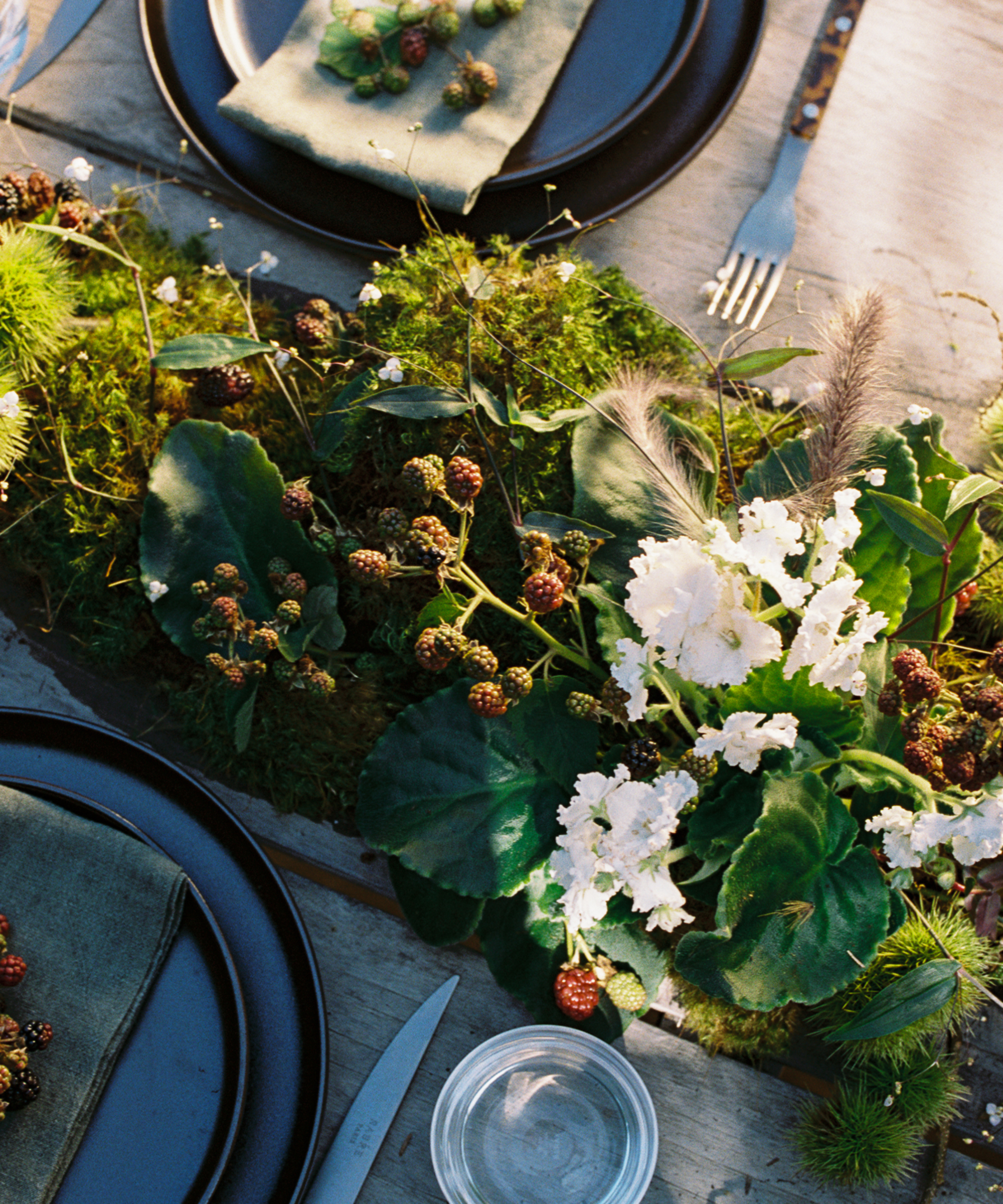 A rustic table setting features green napkins with berries on dark plates, and a lush centerpiece made of moss, green leaves, white flowers, and small clusters of berries. A fork, knife, and glass sit nearby.