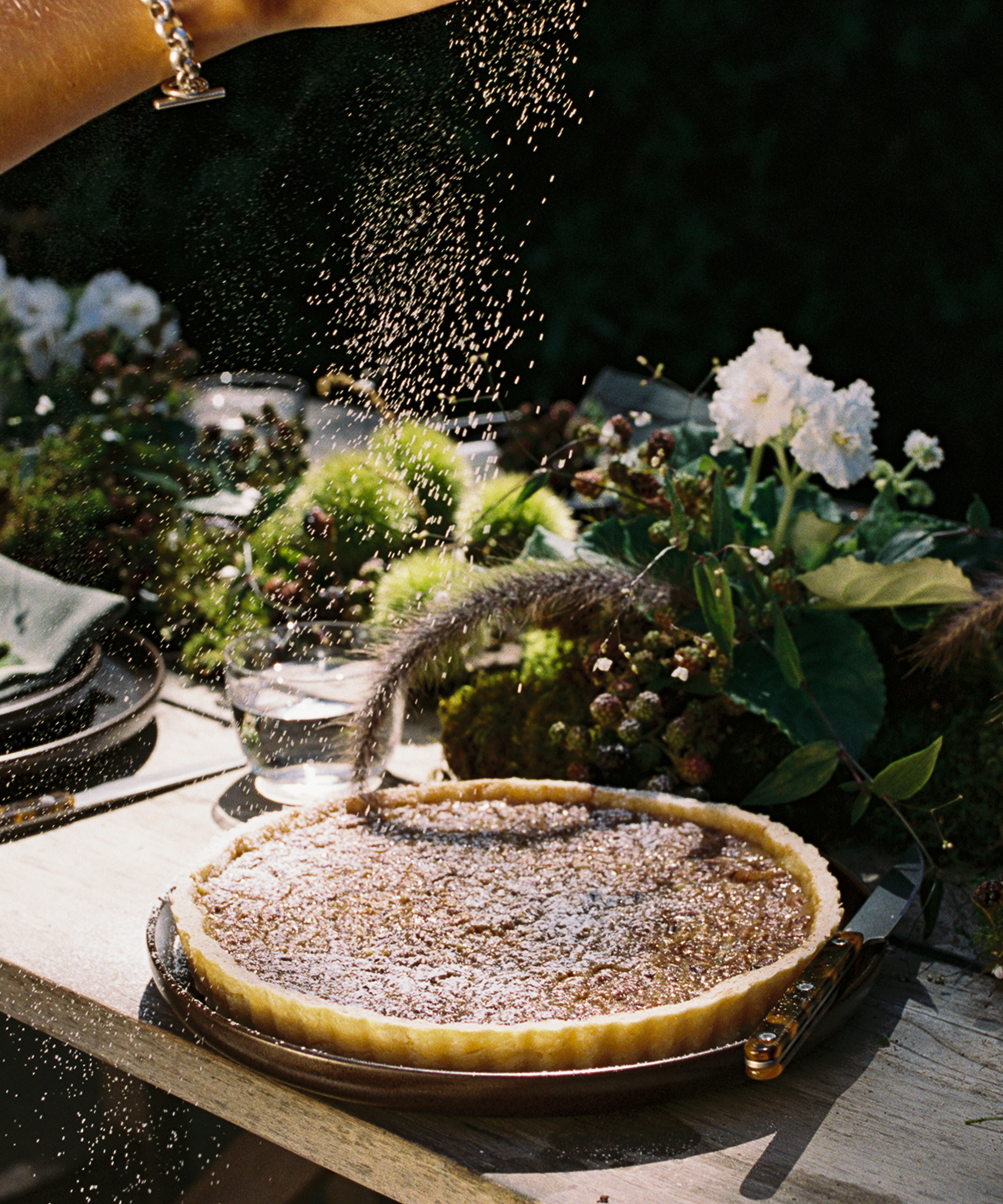 A hand sprinkles powdered sugar over a tart on a wooden table set with flowers, moss, and glassware in natural sunlight.