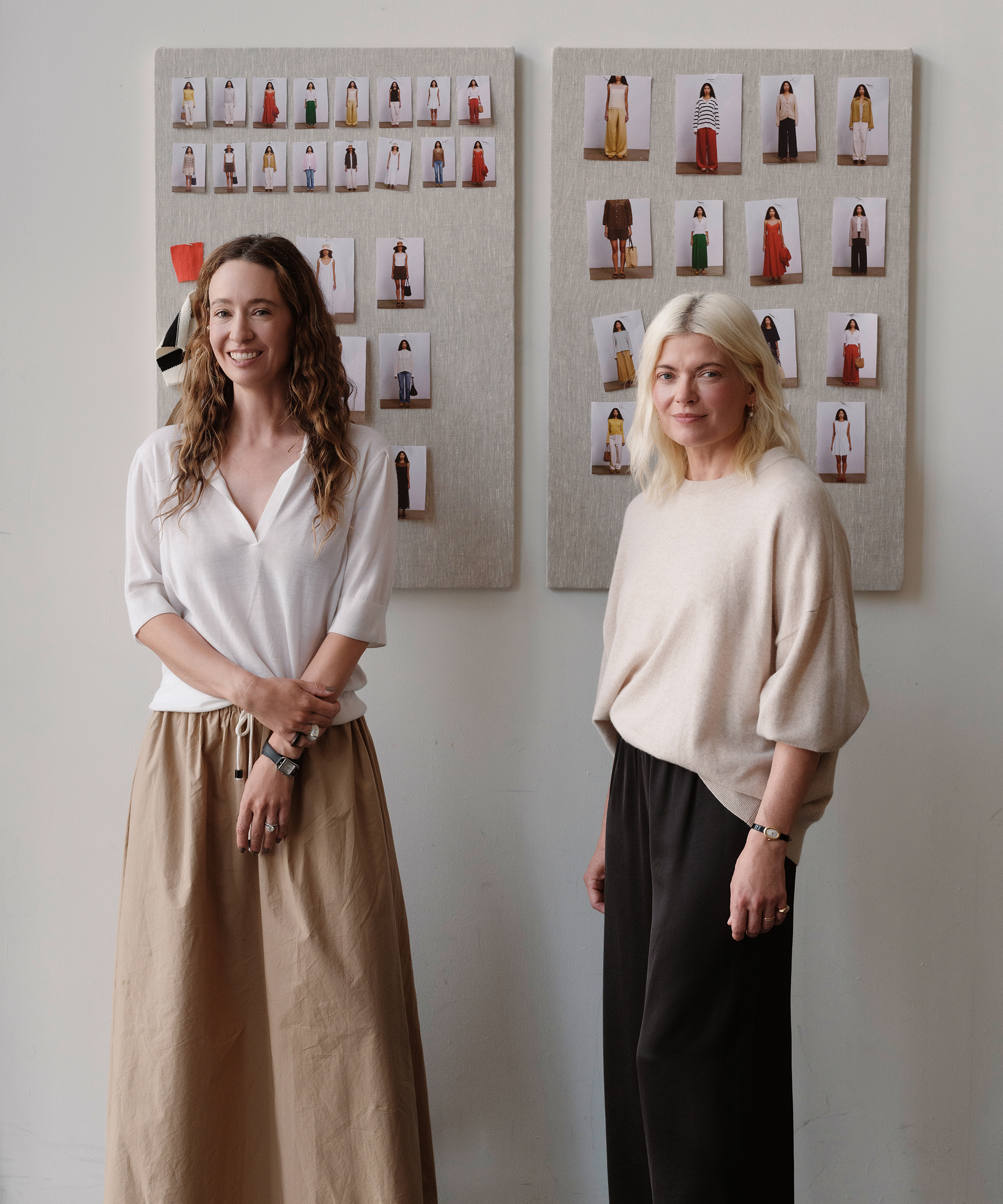Two women stand and smile in front of boards displaying various fashion outfit photos, evoking the spirit of New York Fashion Week. One wears a white blouse and tan skirt, the other a beige top and black pants. The background is minimalist and bright.