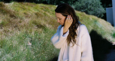 A woman with long brown hair wearing a loose, light-colored sweater from her fall wardrobe walks outside, touching her hair, with green grass and trees in the background.