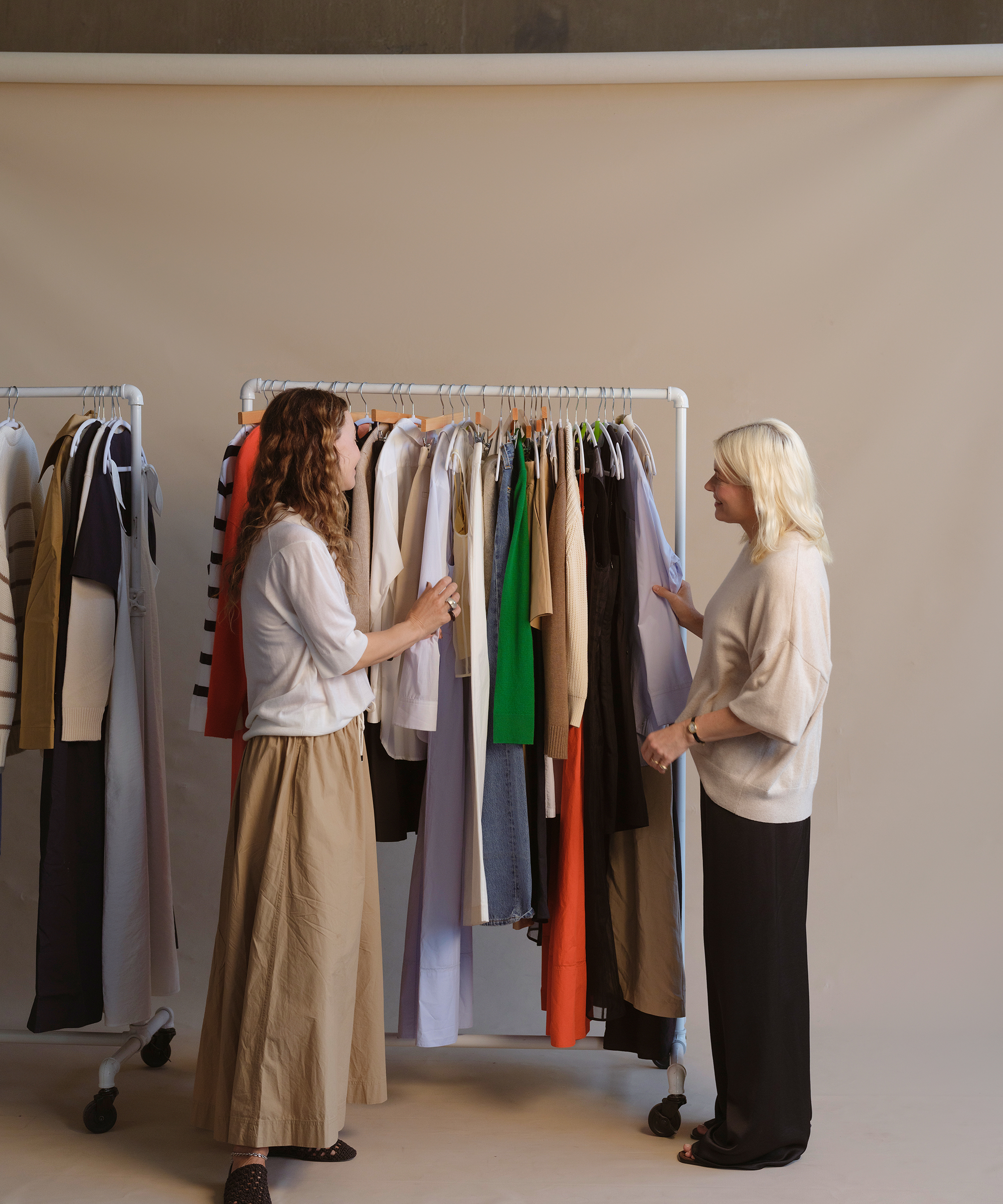 Two women stand facing each other beside clothing racks filled with assorted garments. One woman is selecting a shirt from the rack while the other watches. The background is plain and neutral.