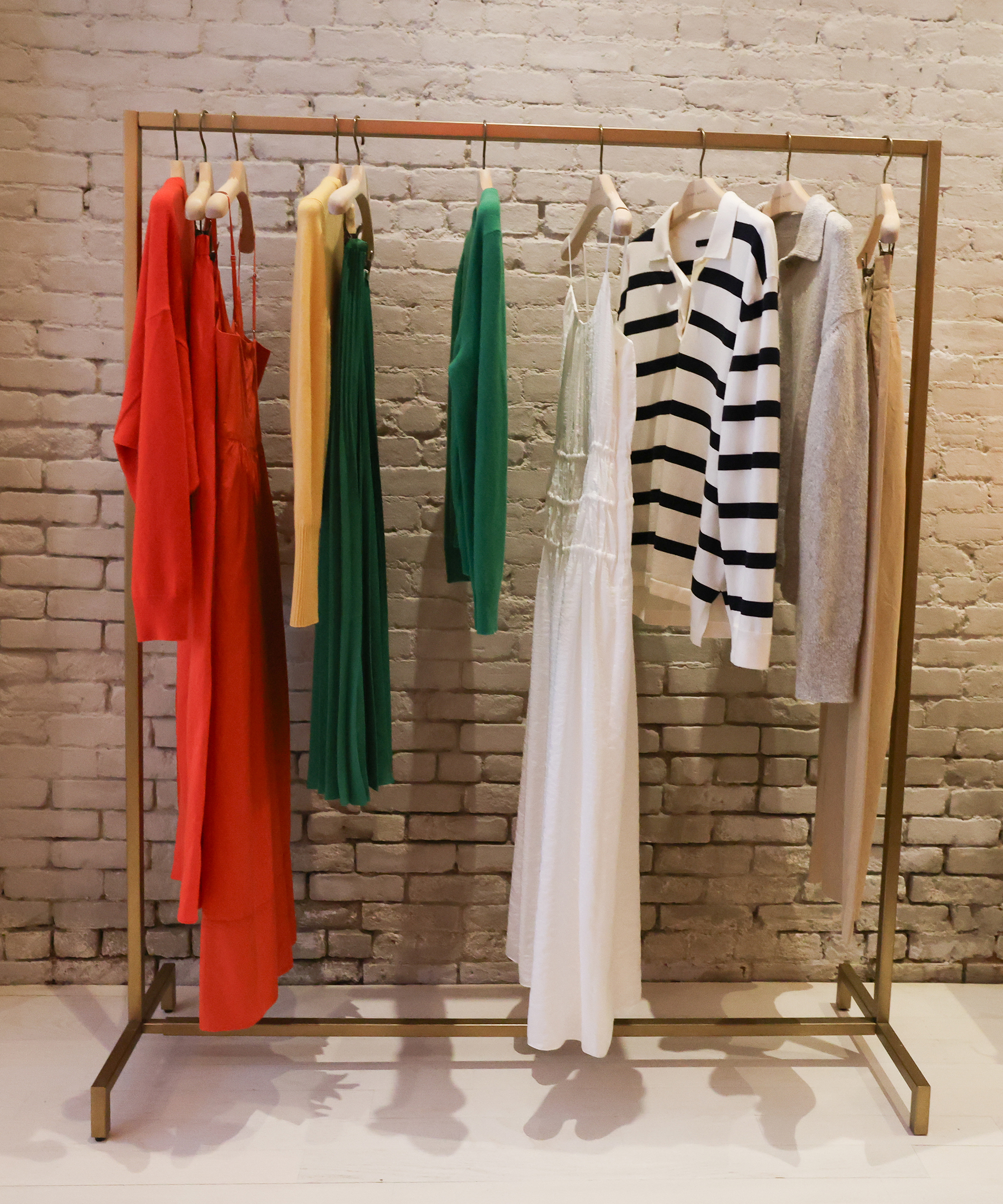 A clothing rack displays various garments, including a red dress, green pants, a yellow sweater, a green sweater, a white dress, a black-and-white striped sweater, a gray sweater, and beige pants against a white brick wall.