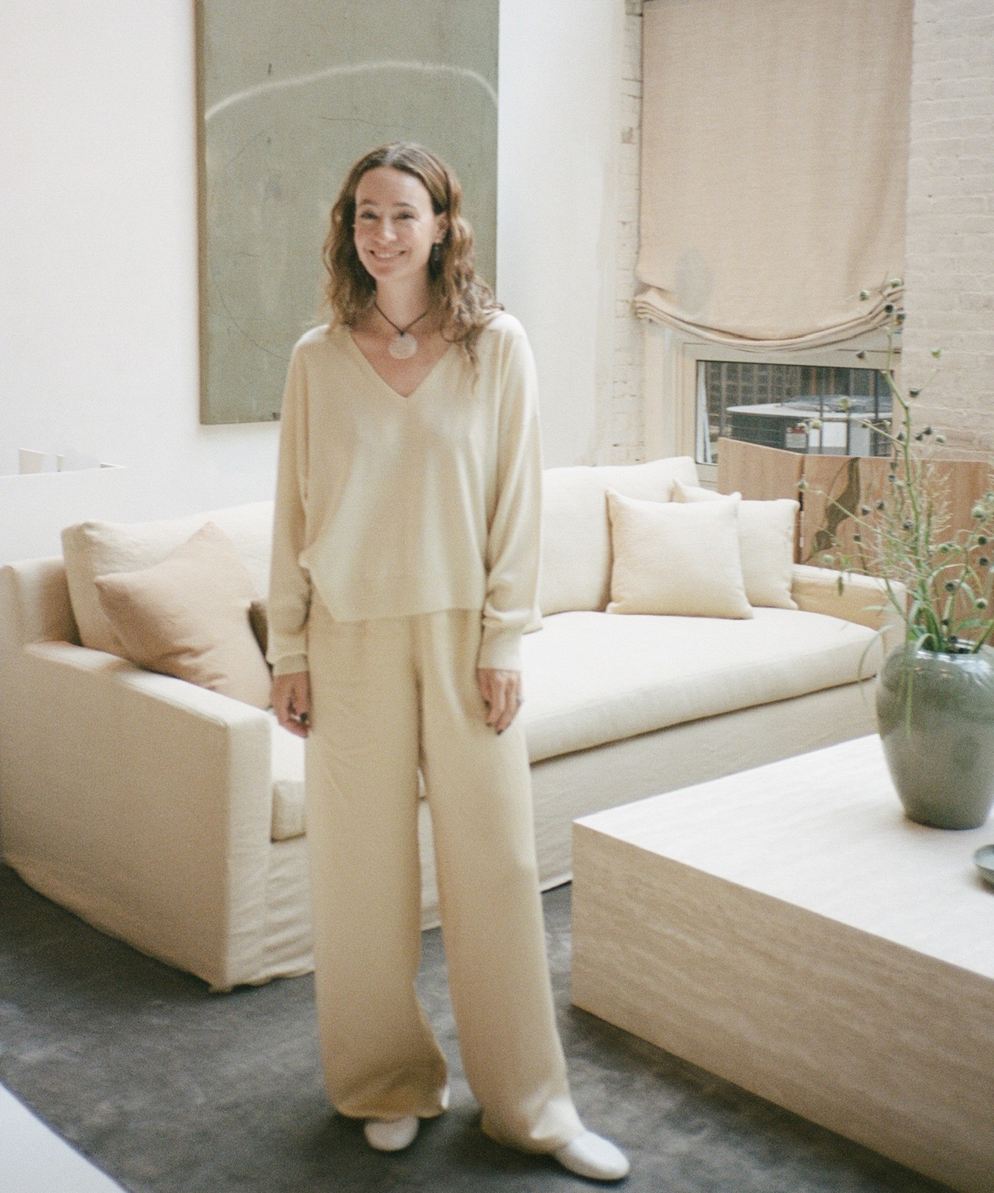 A woman with wavy hair, wearing a loose beige sweater and matching pants inspired by New York Fashion Week, stands smiling in a light-filled modern living room with neutral-toned furniture and minimalist decor.