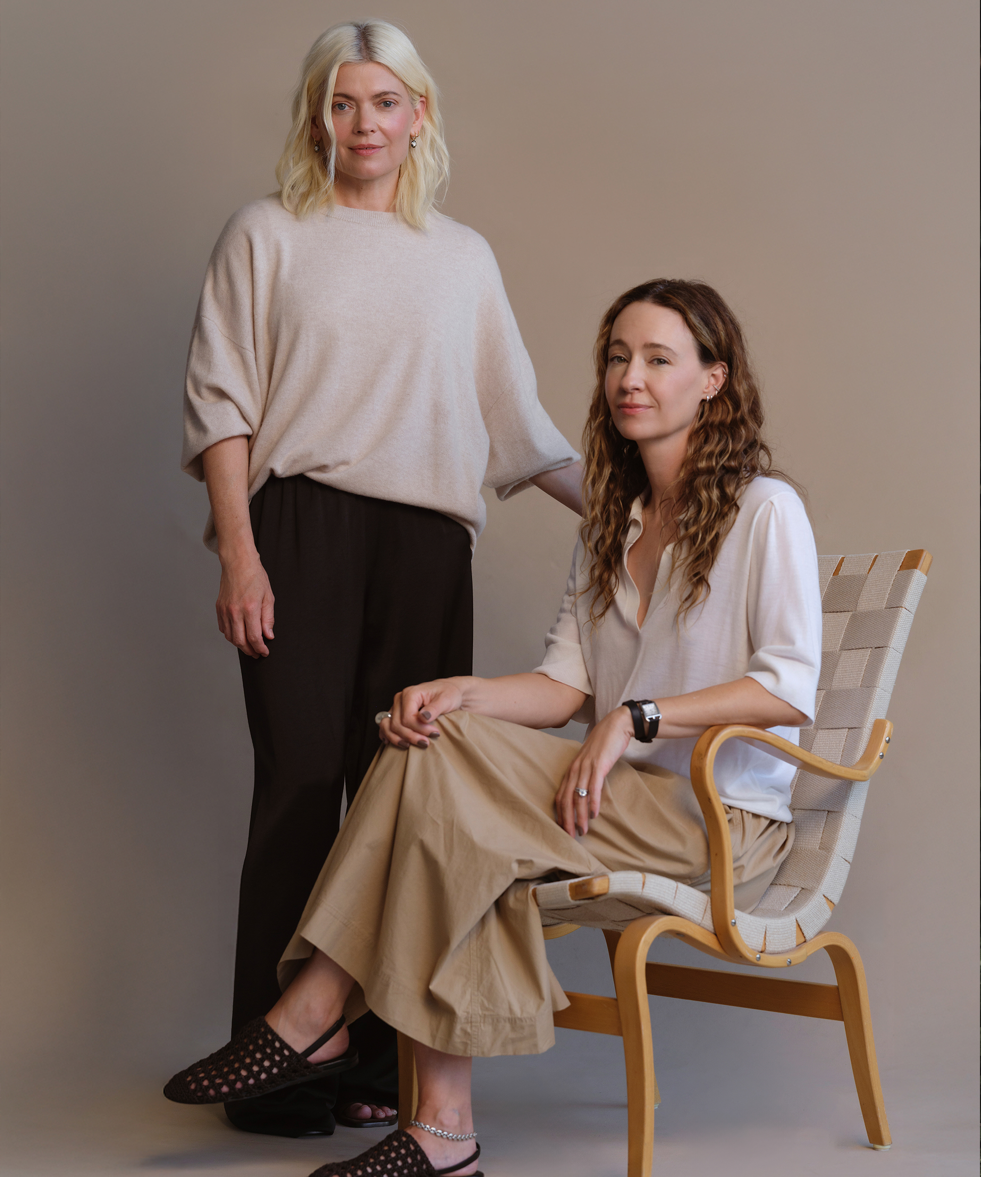 Two women pose for a portrait against a neutral background. One stands wearing a beige sweater and black pants, while the other sits on a chair wearing a white blouse and tan skirt. Both have light-colored hair and calm expressions.
