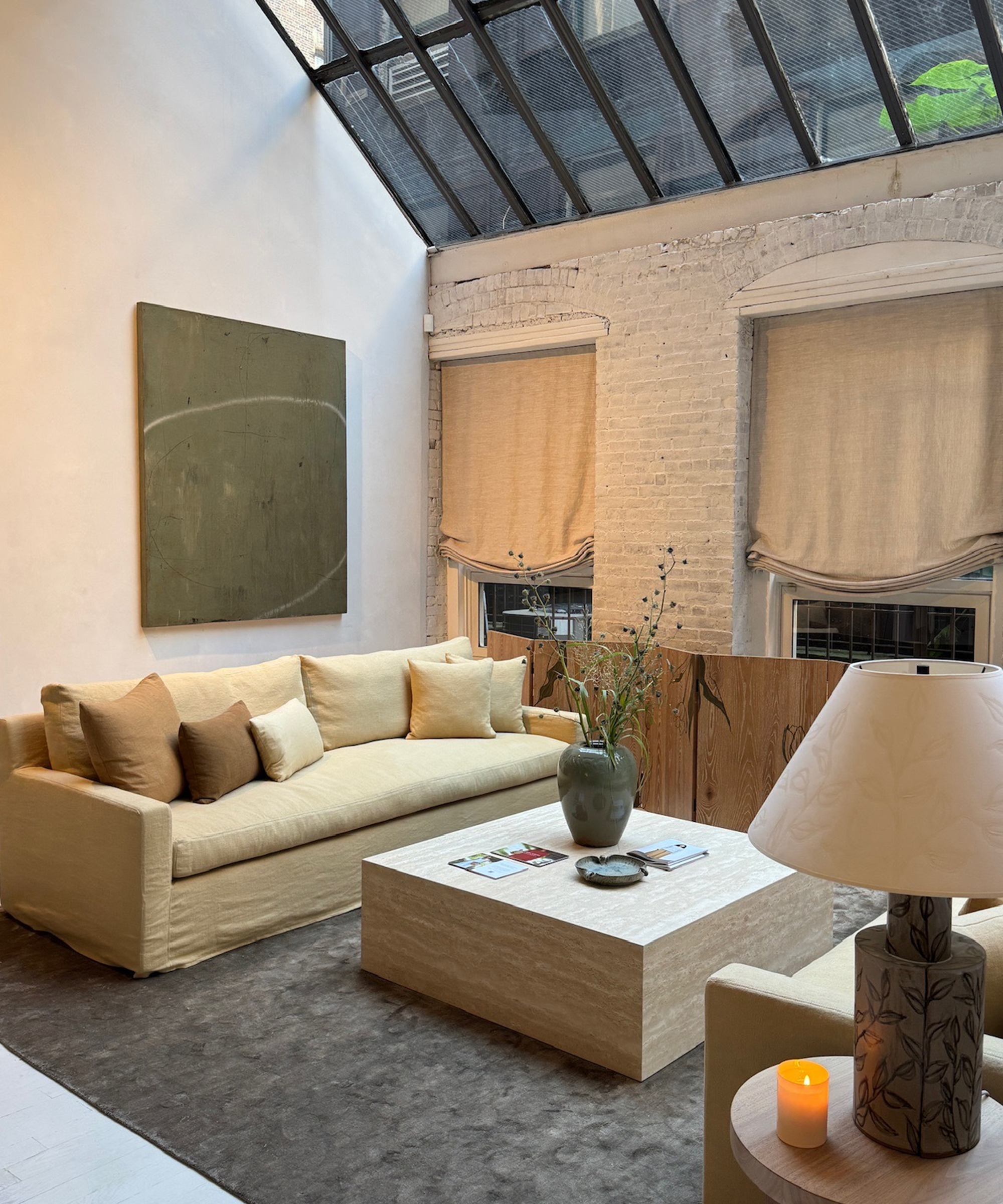 A modern living room with a beige sofa, minimalist art on a white brick wall, and a large coffee table with a vase exudes the chic style of New York Fashion Week. Two windows with beige shades and a glowing lamp sit under a stunning glass ceiling.