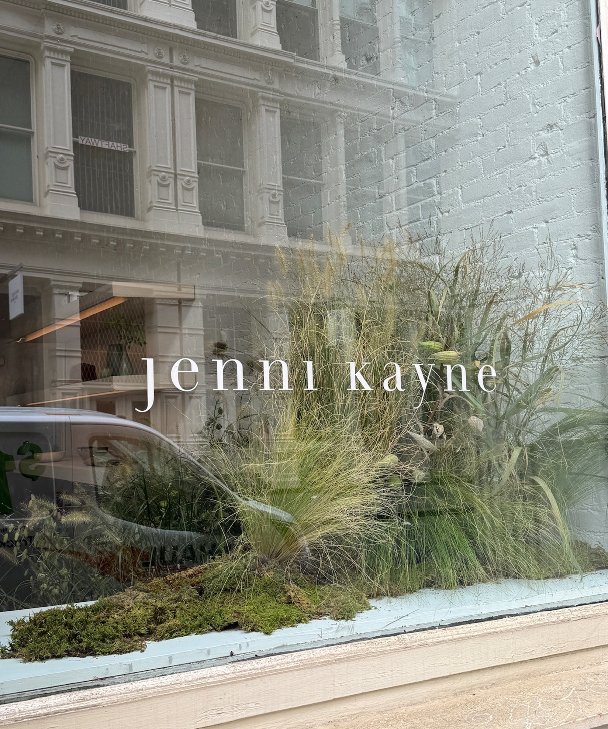 A storefront window displays the words jenni kayne in white text, with green plants and grasses arranged behind the glass—an elegant scene that could fit right into New York Fashion Week, reflecting buildings and a parked car on the street outside.