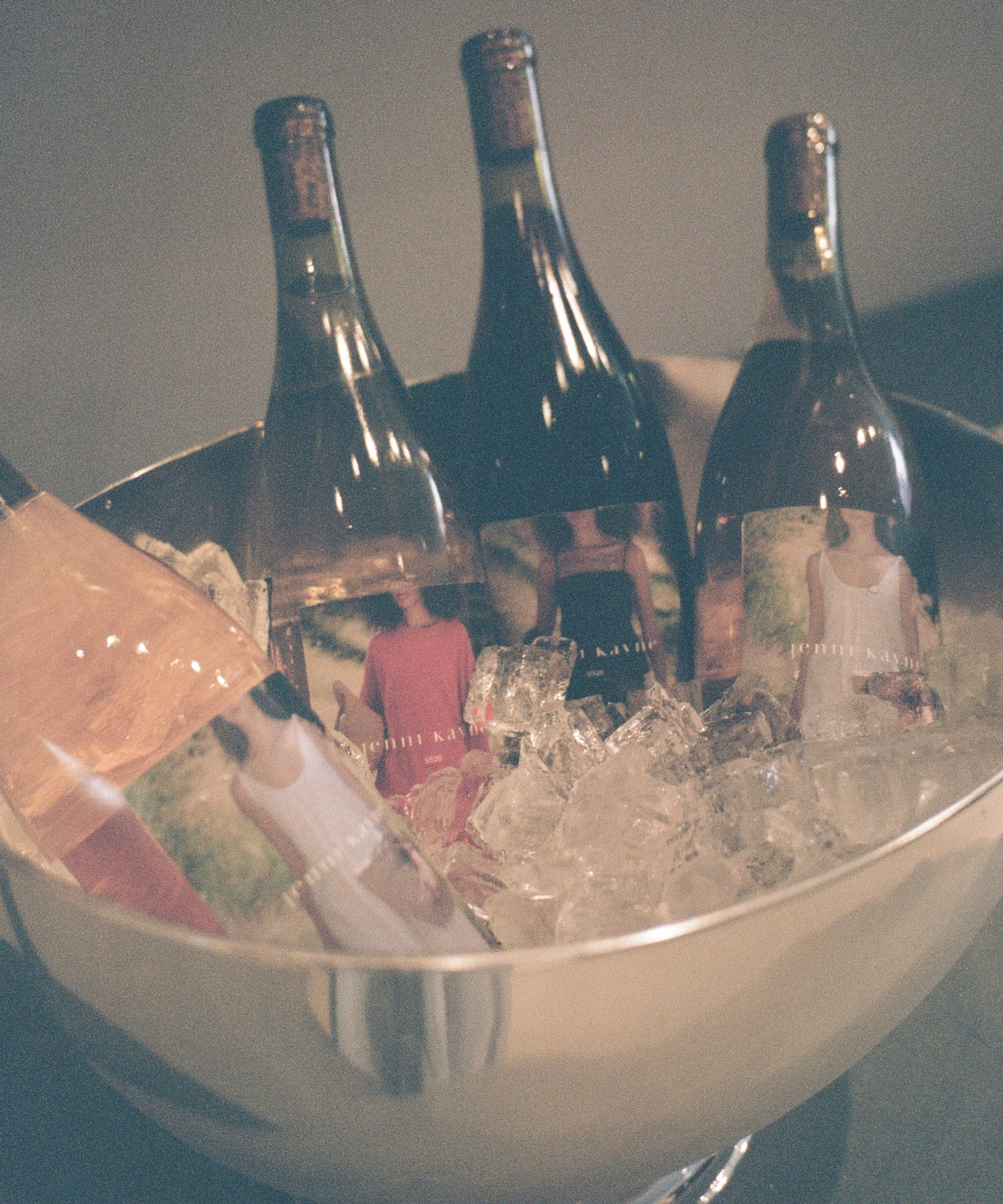 Four wine bottles with colorful labels are chilling in a large silver bowl of ice. The bottles, dressed in outfits inspired by New York Fashion Week, stand out under the soft, muted lighting.