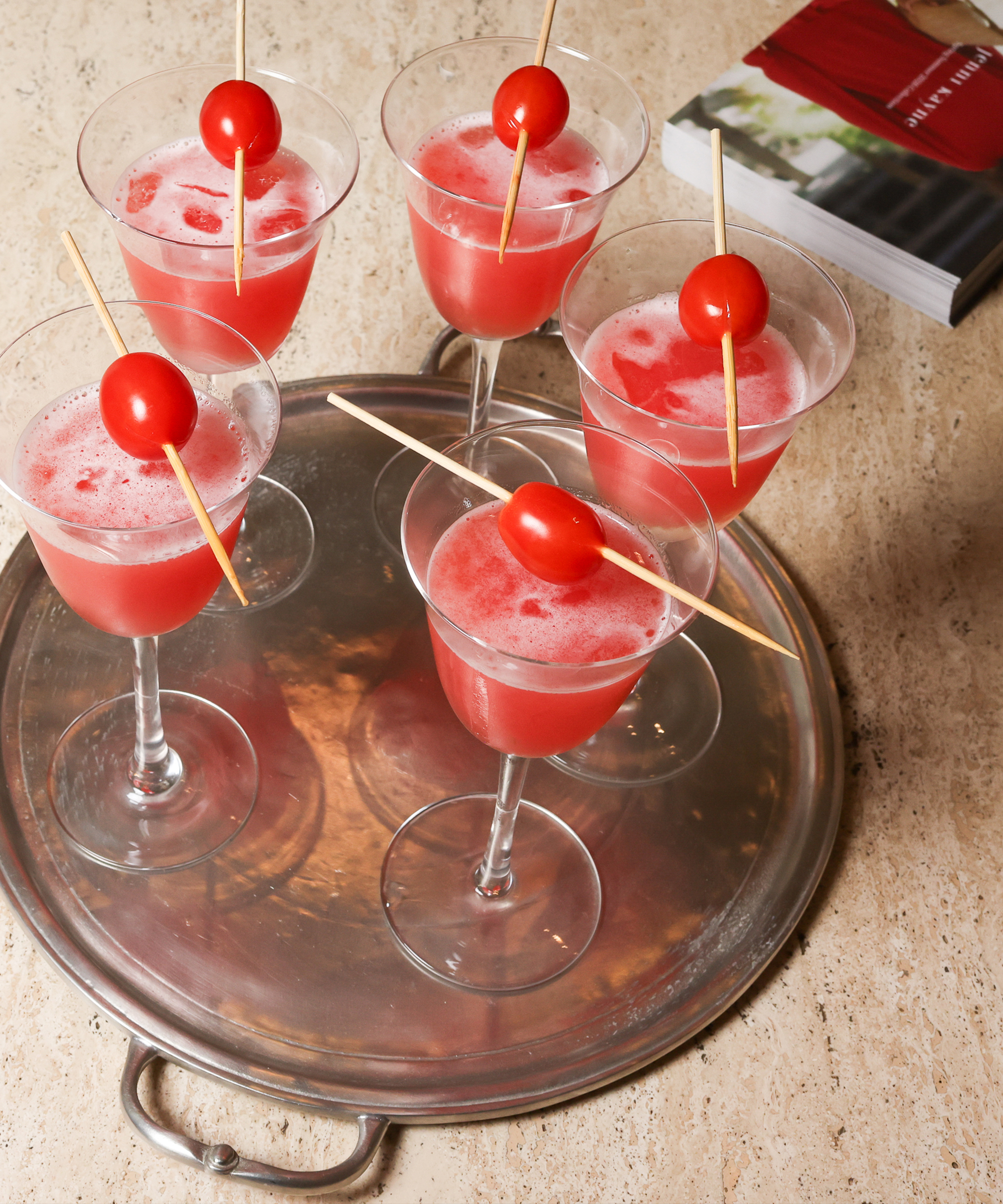 Six stemmed glasses with pink cocktails, each garnished with a red cherry tomato on a wooden skewer, are arranged on a round silver tray. A closed book lies in the background on a beige surface.