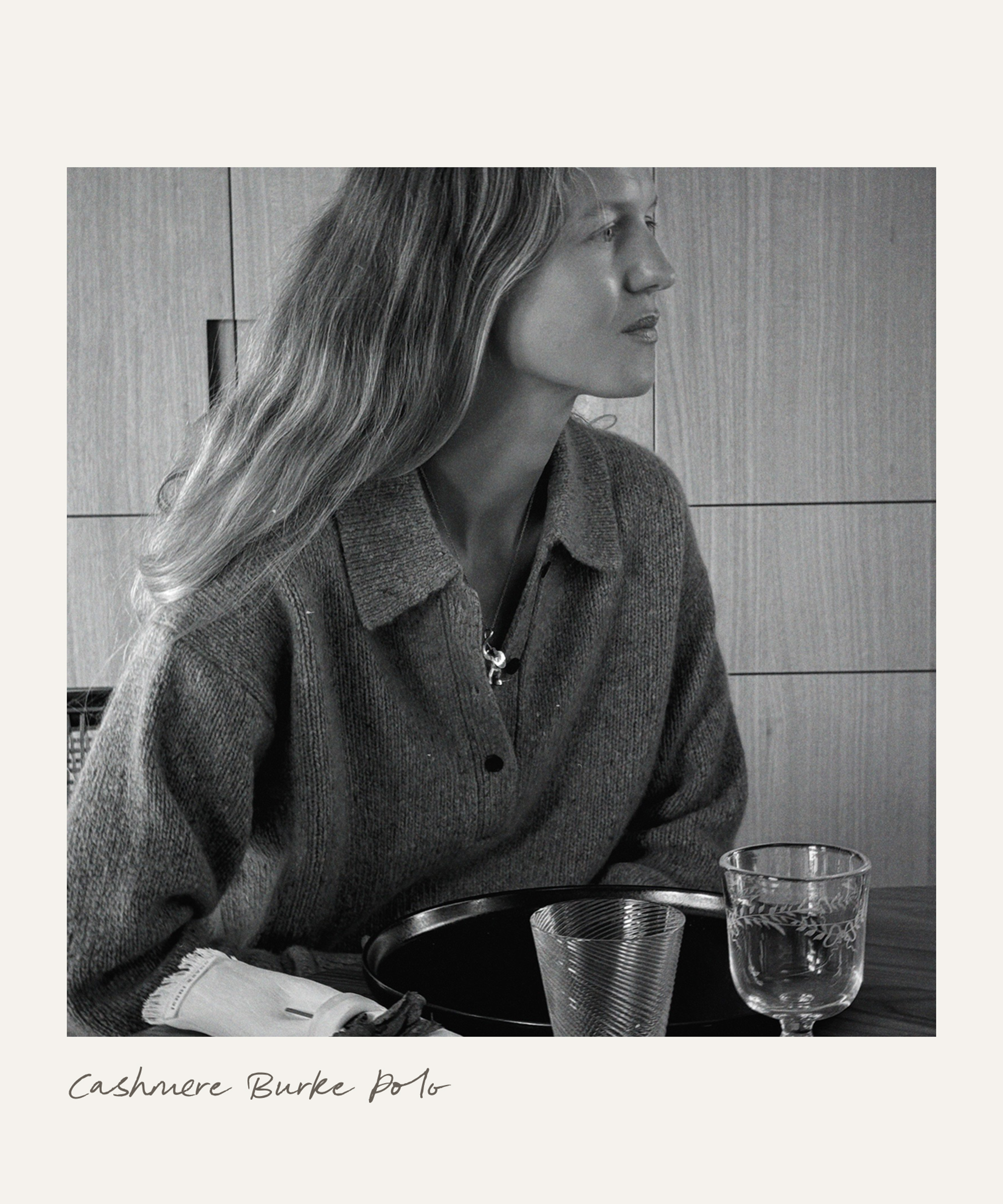 A woman with long hair sits at a table, wearing a buttoned cashmere polo sweater. Two glasses and folded napkins are in front of her. The image is in black and white, with Cashmere Burke Polo written below.