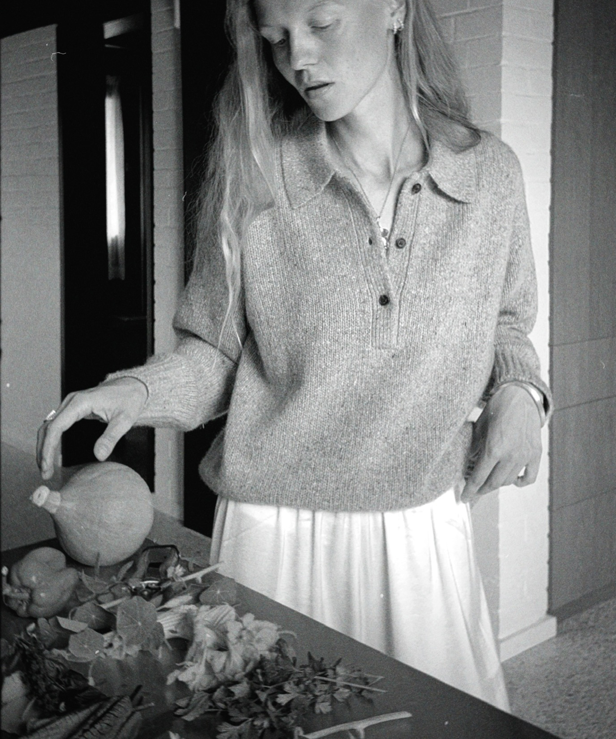 A woman with long hair and a light sweater stands beside a table, arranging various vegetables, leaves, and herbs. The setting appears to be indoors, and the photo is in black and white.