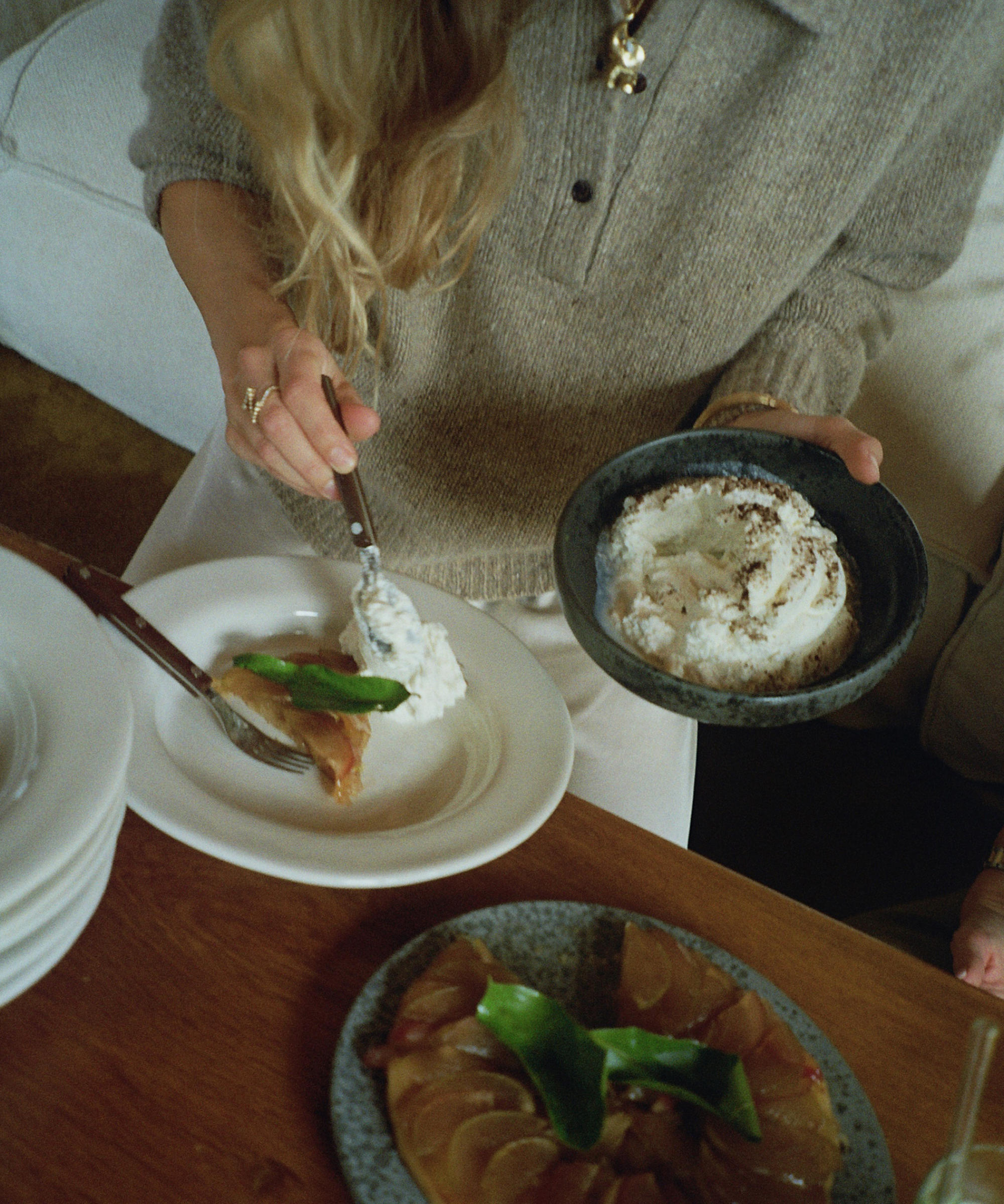 A person with long blonde hair serves a dessert, possibly cake with whipped cream, onto a white plate. They hold a bowl of cream and sit at a wooden table set with plates of food.