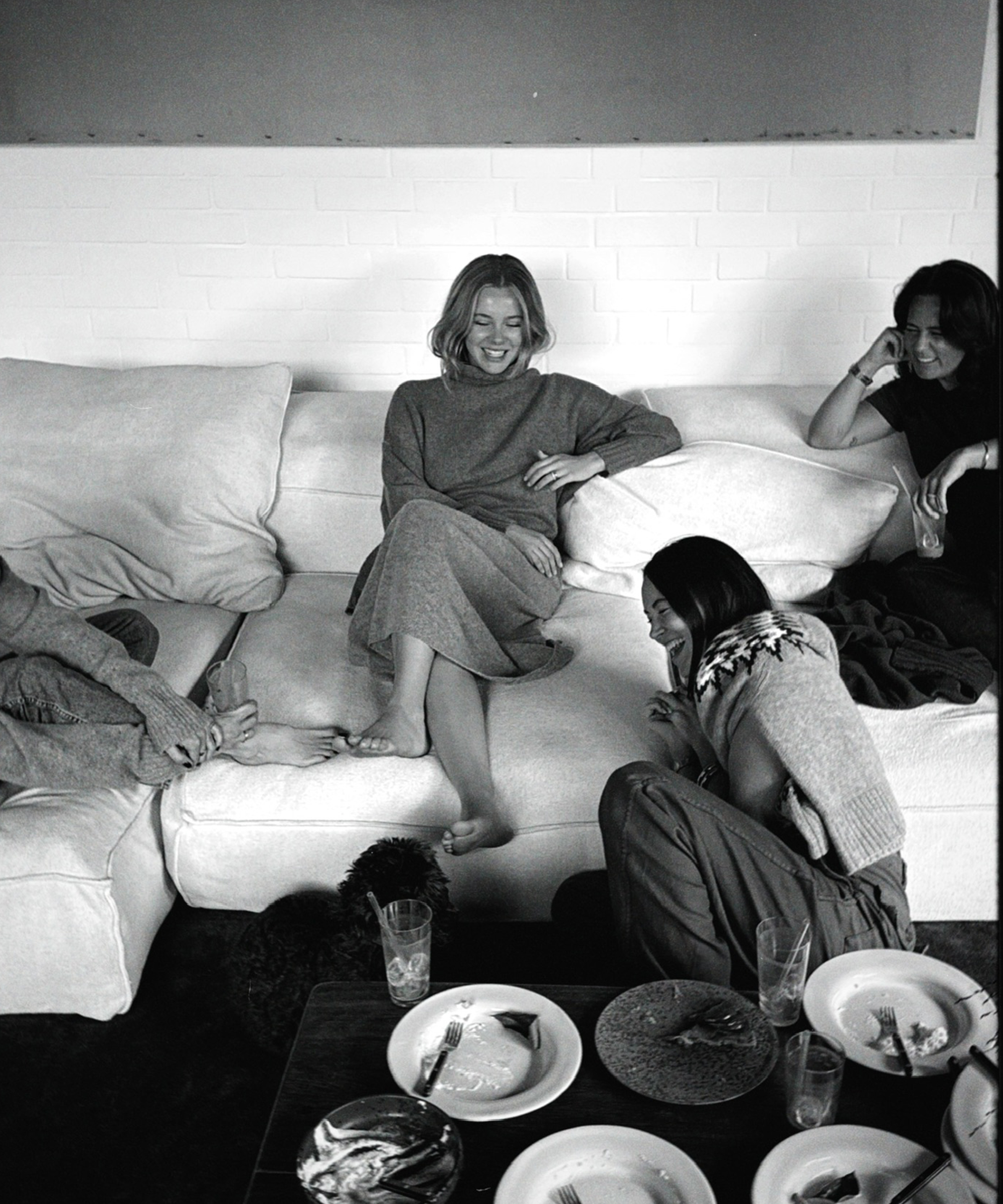 Four people relax on a white couch, smiling and chatting. Plates with leftover food and drinks are on a table in front of them. A small dog sits on the floor. The scene feels casual and friendly.