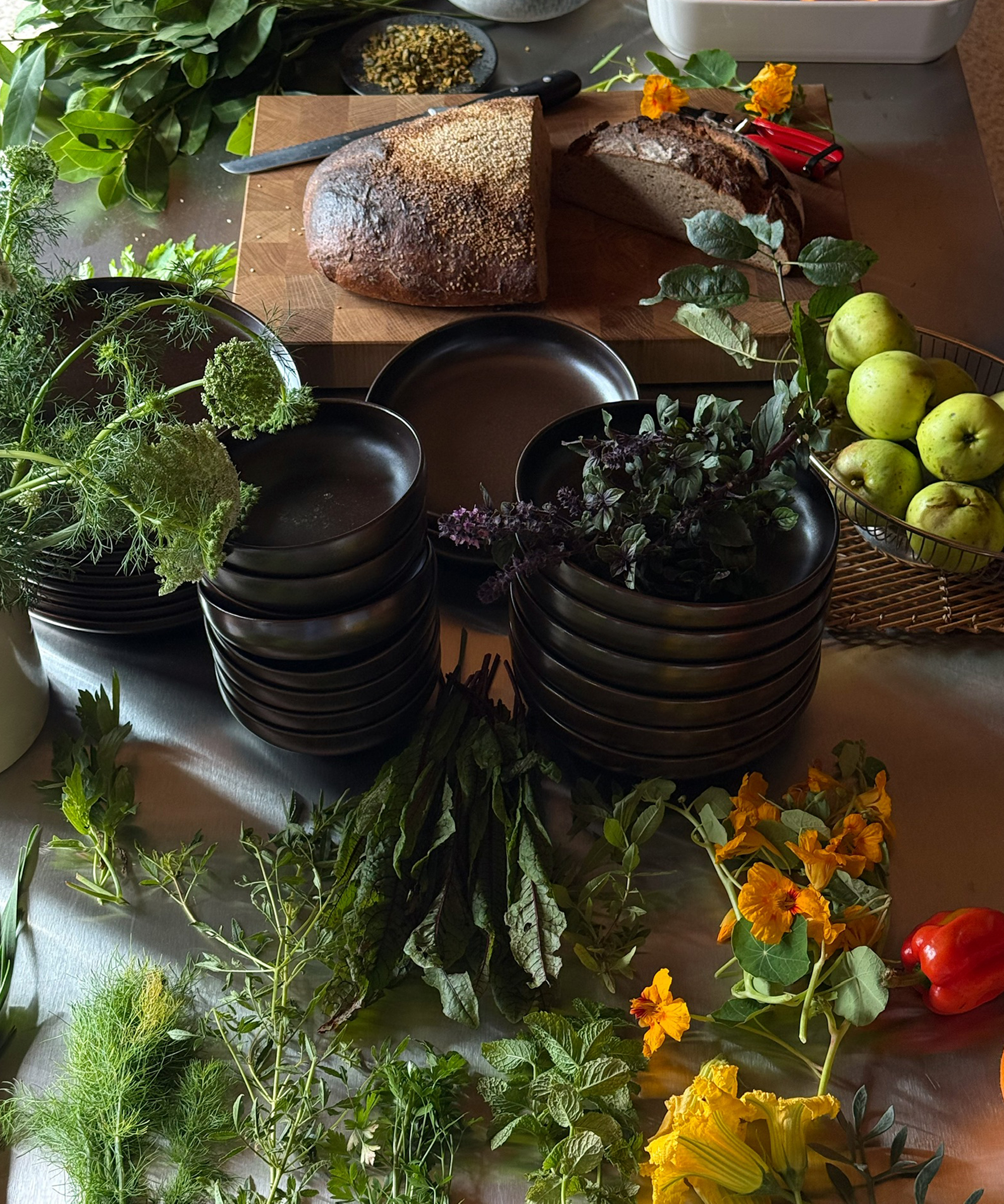 A table with stacks of black plates, a sliced loaf of bread, green apples in a basket, assorted fresh herbs, leafy greens, and orange edible flowers arranged around.