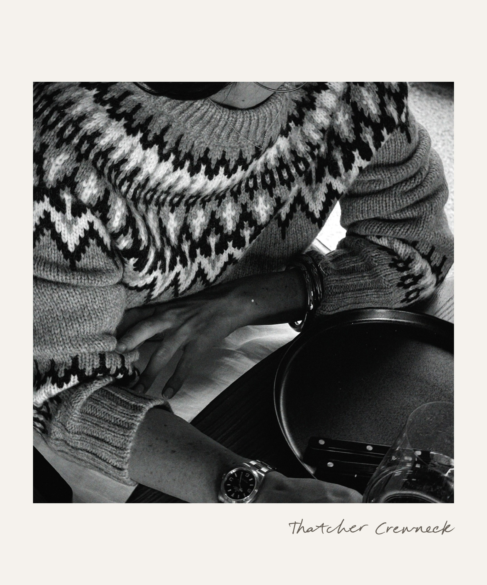 A person in a patterned knit sweater sits at a table with crossed arms, next to a plate, cutlery, and a glass. Only the lower half of their face is visible. The photo is black and white.