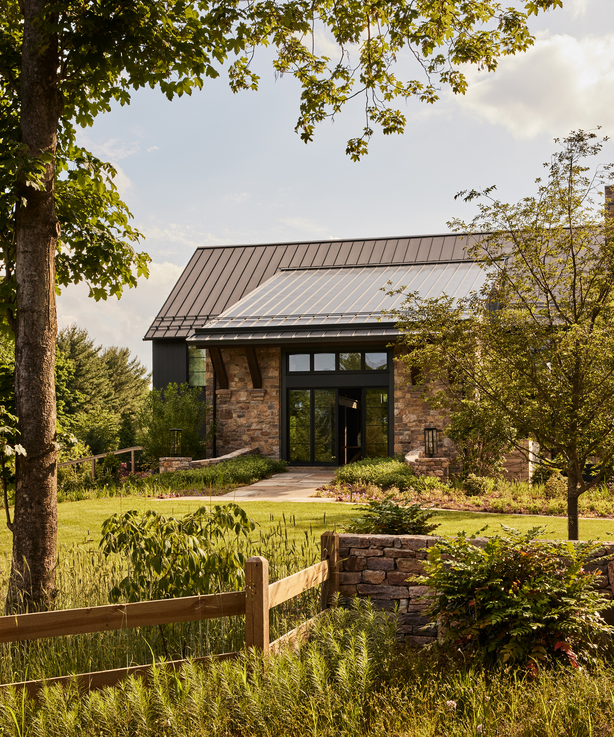 A modern farmhouse with a metal roof and stone exterior—styled by Ursino Interiors—sits amid lush greenery, gardens, and trees, with a wooden fence and stone wall in the foreground on a sunny day.