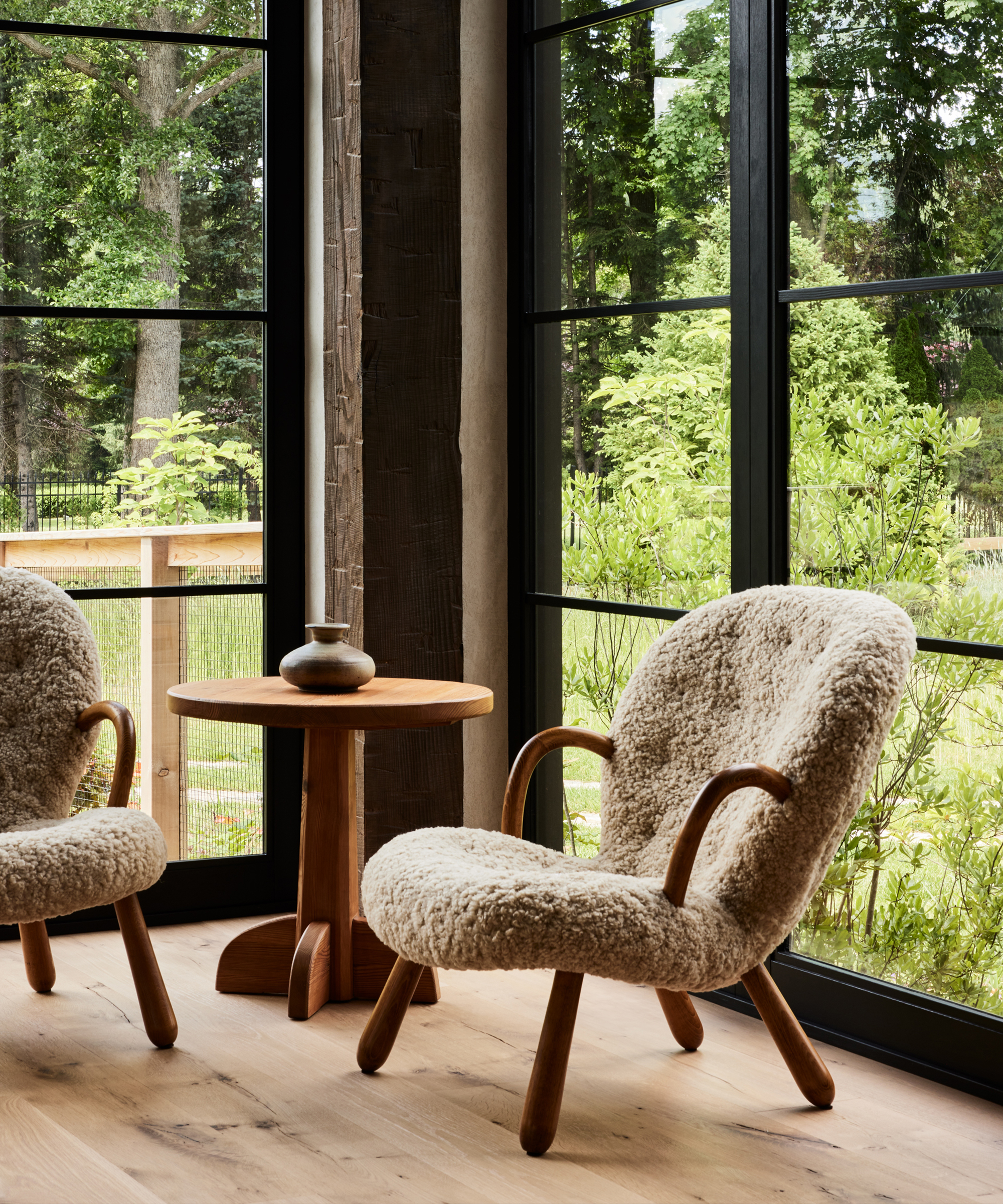 A cozy corner by Ursino Interiors features two plush sheepskin armchairs and a small wooden table with a vase, set by large windows overlooking a lush, green garden.