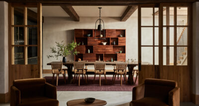 A modern dining room by ursino interiors features a long wooden table, eight woven chairs, a purple rug, and pendant lights. Wooden beams and sliding glass doors frame the space, with two armchairs in the foreground and a large shelf with decor.