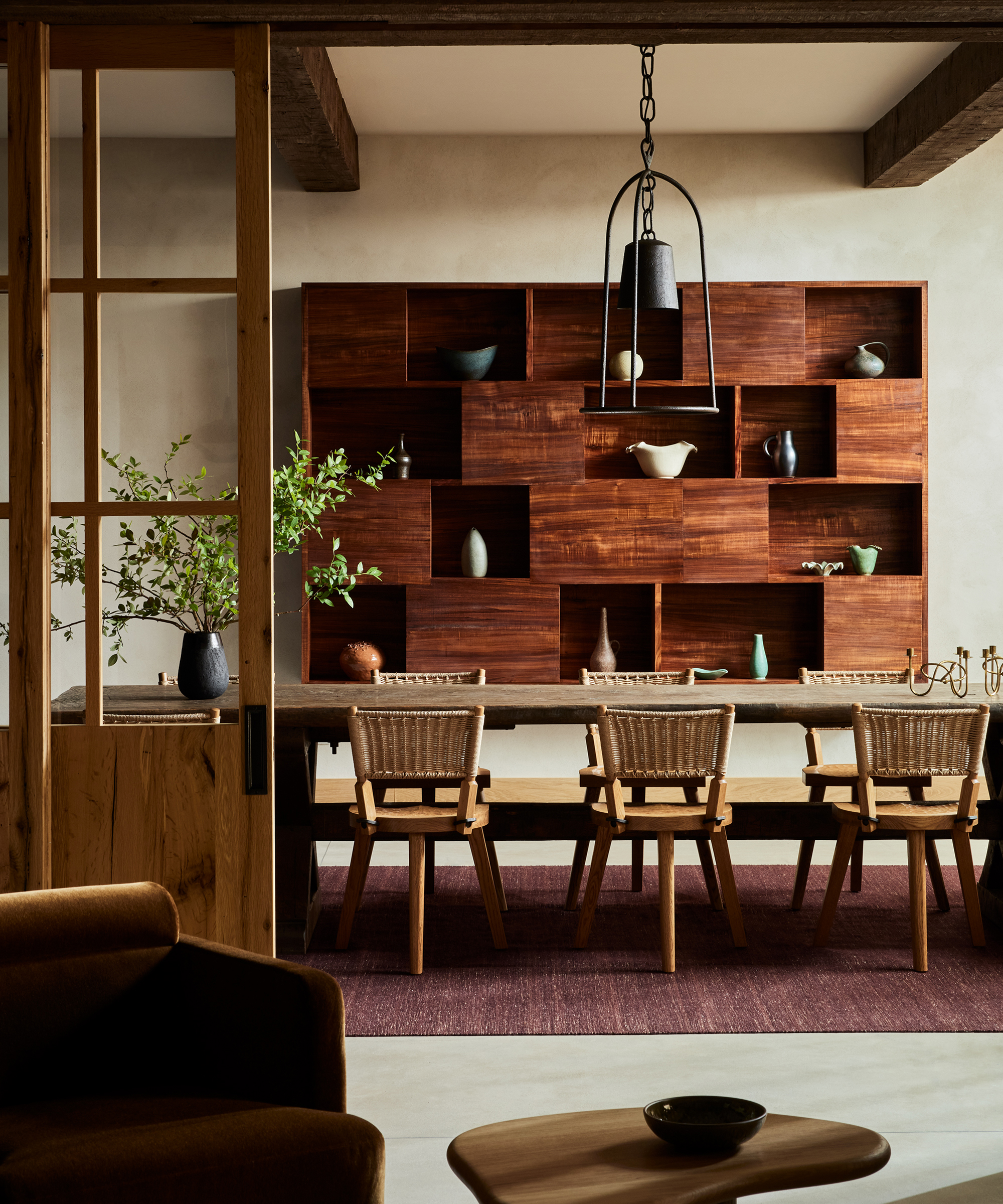 A modern dining room by Ursino Interiors features a wooden table, six woven chairs, a large wooden shelving unit displaying ceramics, a pendant light, a vase with branches, and a muted color palette.