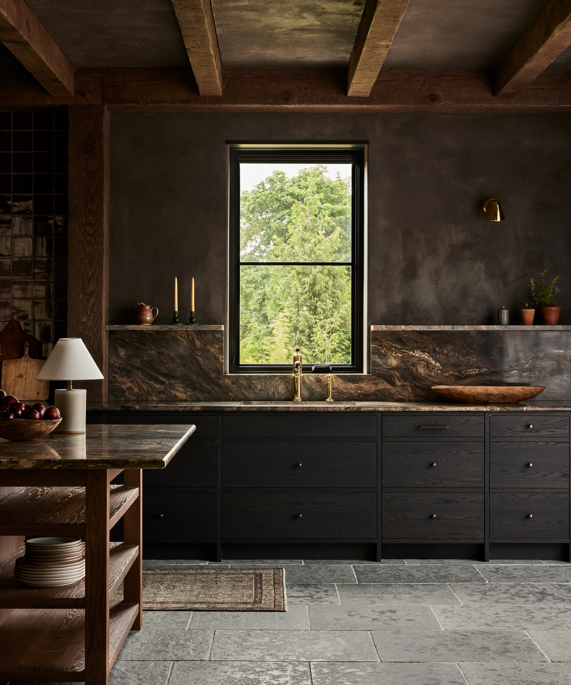 A rustic kitchen by Ursino Interiors features dark wood cabinets, stone countertops, and a central window showcasing greenery. Exposed wooden beams and minimalist decor like a table lamp, candles, and potted plants complete the look.