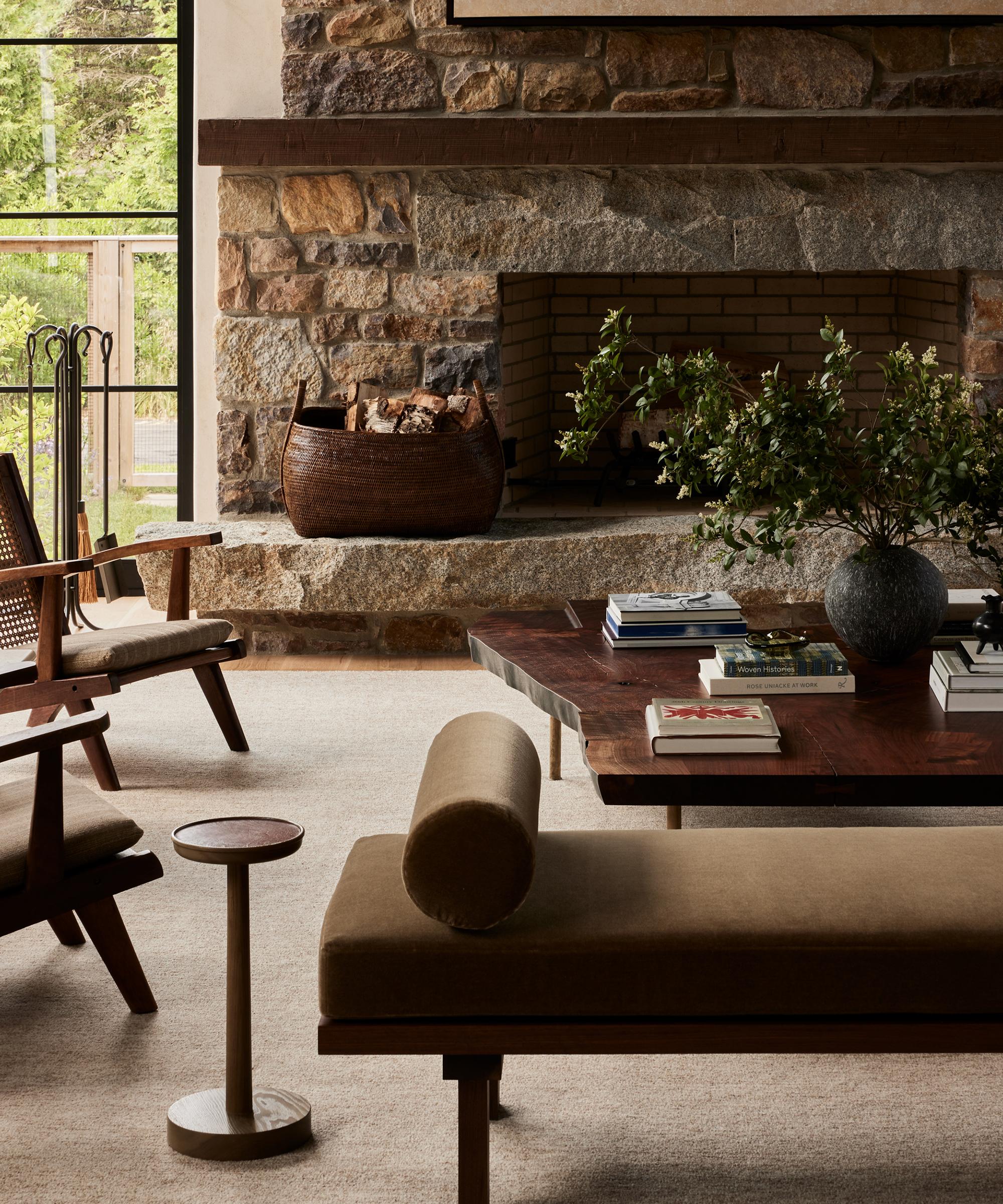 A cozy living room by ursino interiors with a stone fireplace, wooden chairs, a brown bench, and a low table topped with books and greenery. Large windows frame green trees, while earthy tones and natural textures complete the inviting decor.