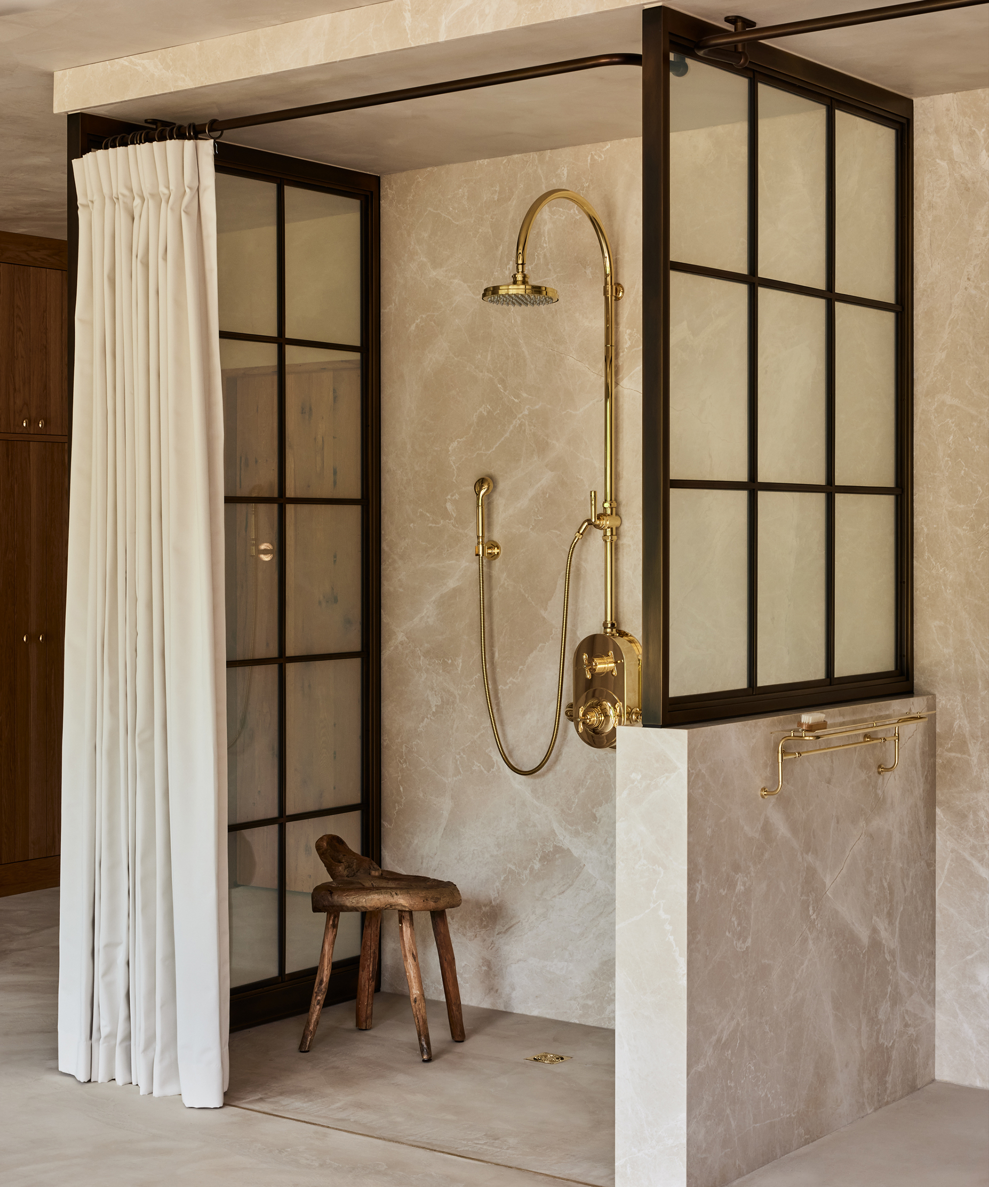 A modern shower by Ursino Interiors features brass fixtures, a rainfall showerhead, glass panels with black grid frames, a white curtain, beige marble walls, and a rustic wooden stool.