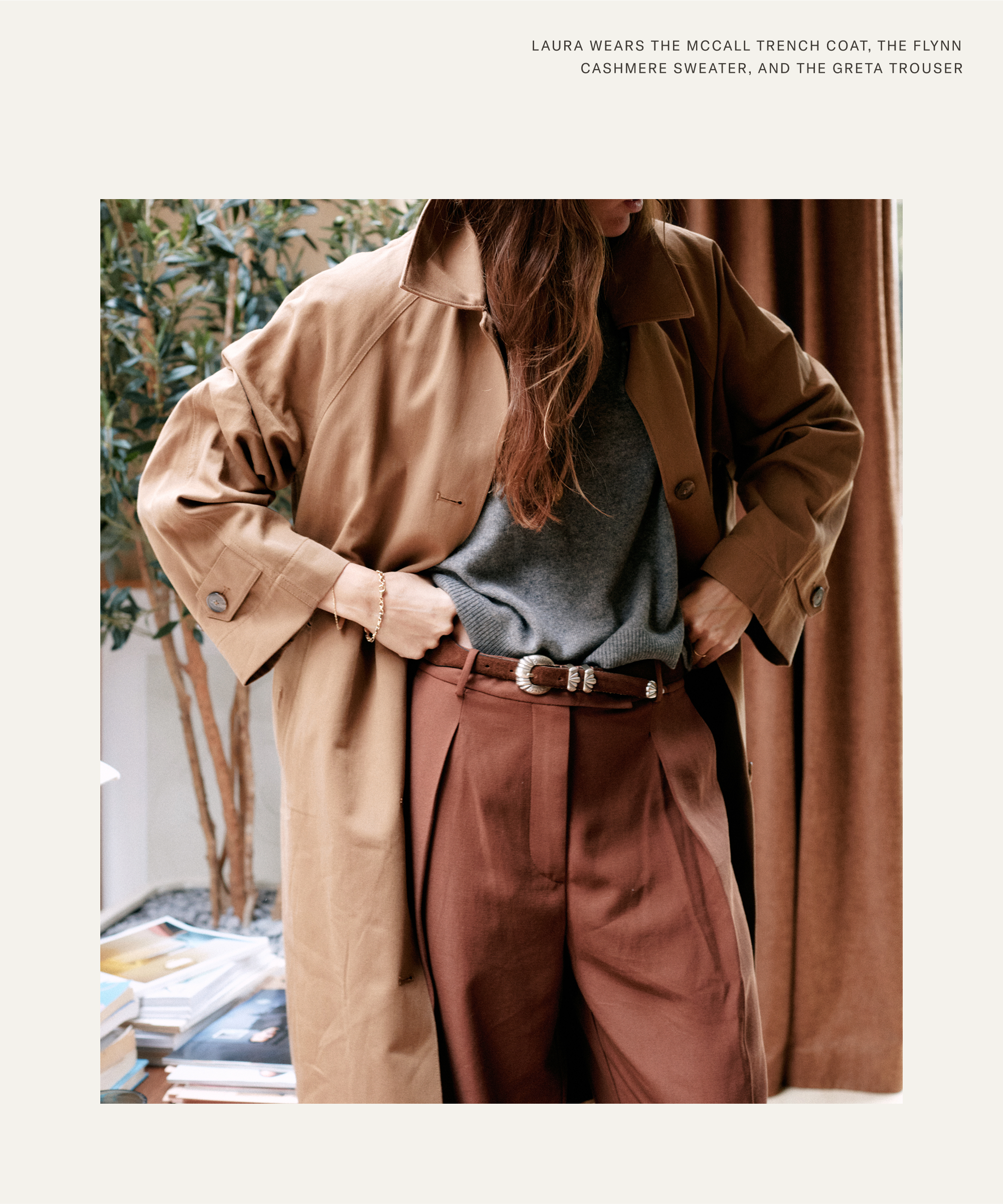 A woman stands indoors wearing a tan trench coat over a gray sweater and brown trousers, channeling Laura Vidrequin Roso’s effortlessly chic style. One hand rests on her hip as her brown hair falls partly across her face. A stack of books and a leafy plant sit behind her.