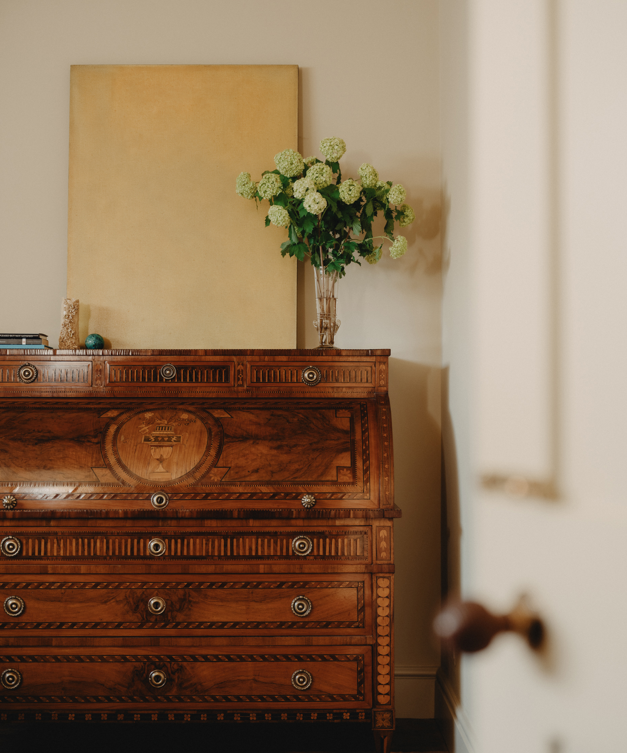 A classic Berkeley-Hawkes wooden chest of drawers with ornate patterns stands against a cream wall. On top are a clear vase of green hydrangeas, a stack of books, and a large blank canvas resting behind them.