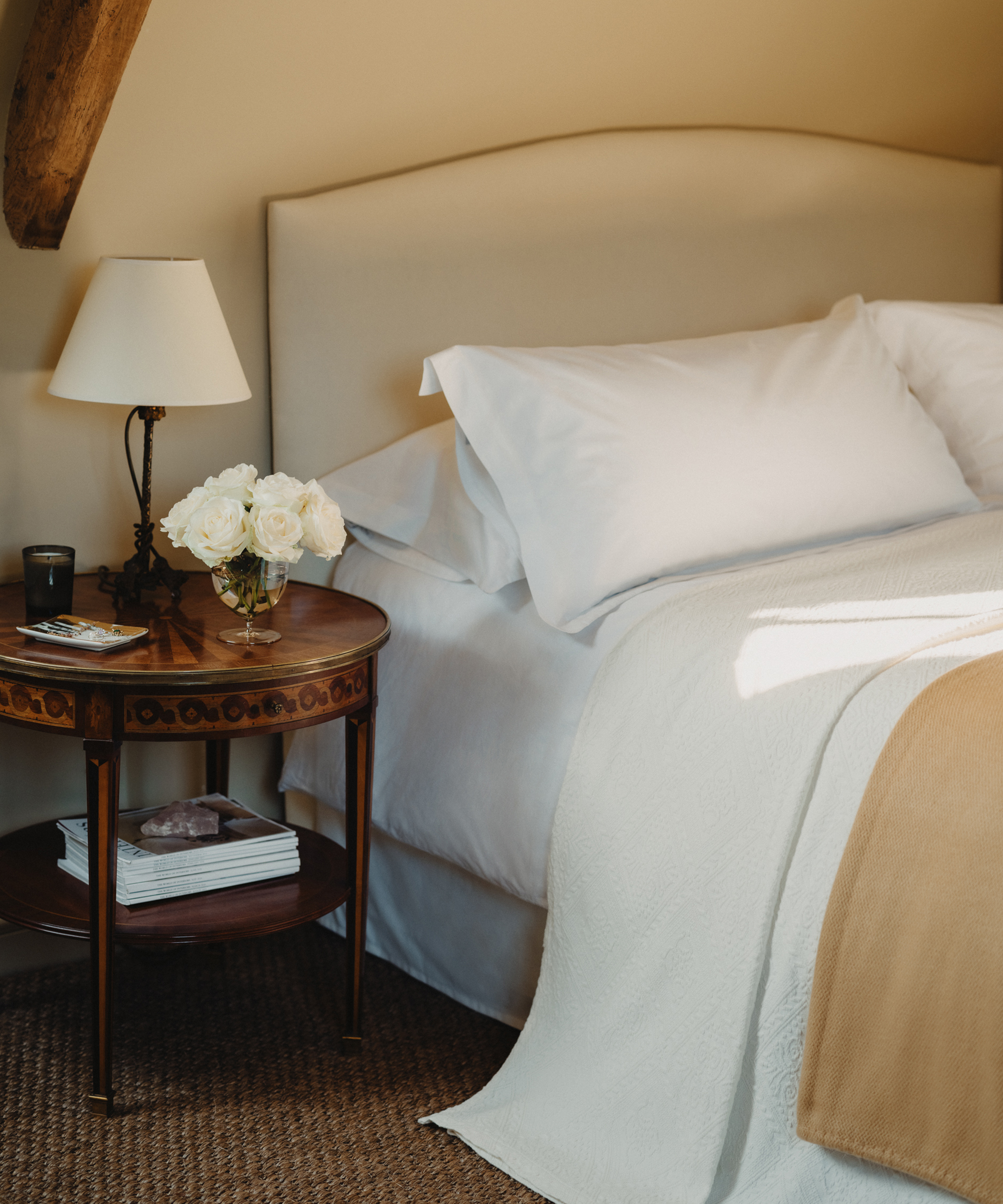 A cozy Berkeley-Hawkes bedroom features a neatly made bed with white bedding, a light brown blanket, and plush pillows. A round wooden nightstand holds a lamp, a vase of white roses, a candle, and stacked magazines below.