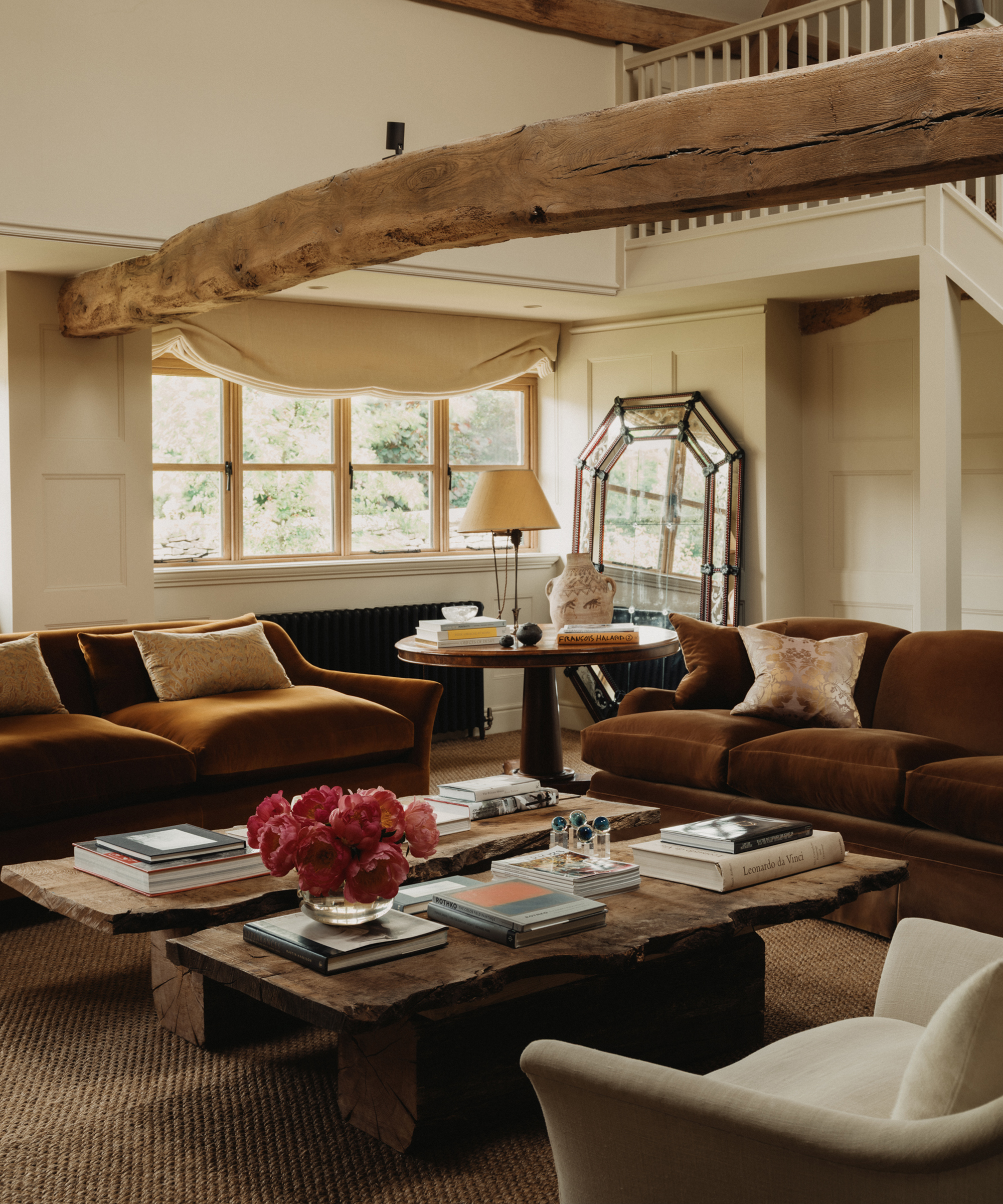 A cozy Berkeley-Hawkes living room with brown velvet sofas, rustic wooden coffee tables, a vase of pink flowers, books, large windows, exposed wooden beams, and a loft area with a white railing.