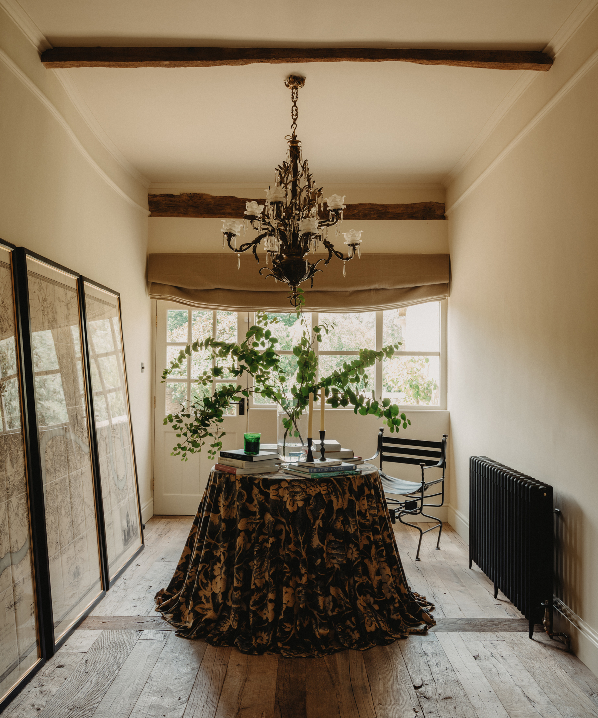 A narrow Berkeley-Hawkes room with wooden beams, large windows, and a decorative chandelier. A round table with a ruffled patterned cloth holds books, vases, and leafy branches. Maps lean against the wall; a black chair and radiator are nearby.