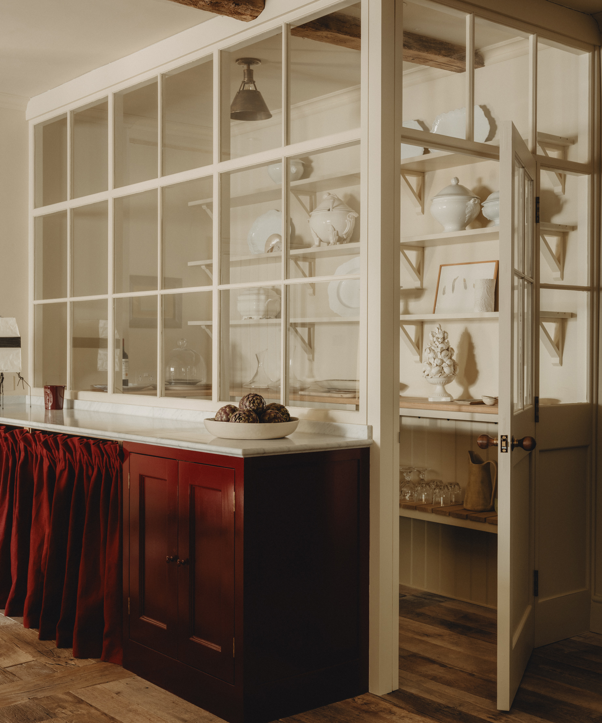 A cozy kitchen with a glass partition, white shelves holding dishes and teapots, a Berkeley-Hawkes red cabinet with a marble counter, and a red curtain below. An open door leads to a pantry with more shelves and glassware.