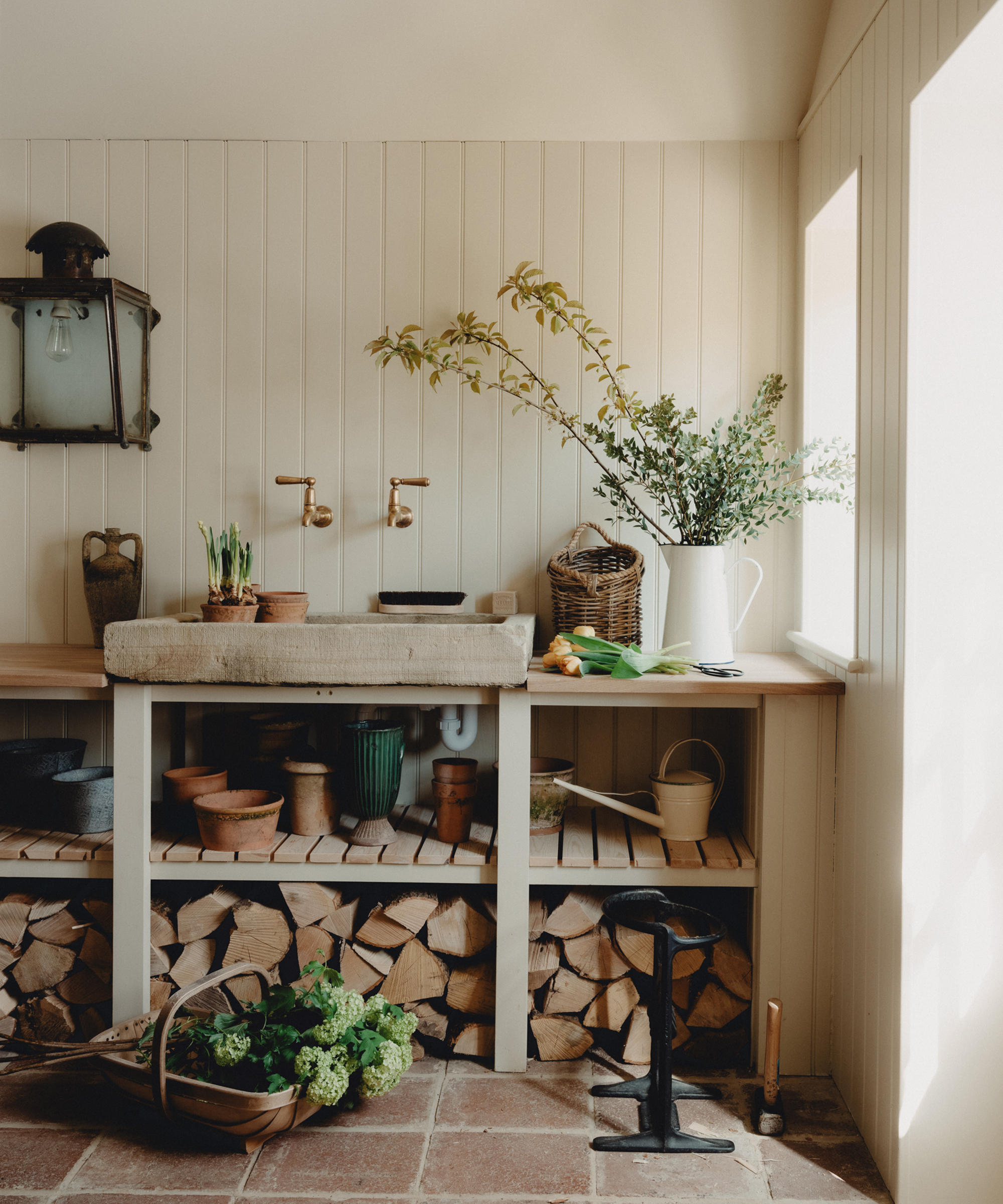 A rustic Berkeley-Hawkes potting bench with a stone sink, brass taps, potted plants, gardening tools, firewood, and a window letting in natural light. A white jug with greenery sits on the counter next to baskets and pots.