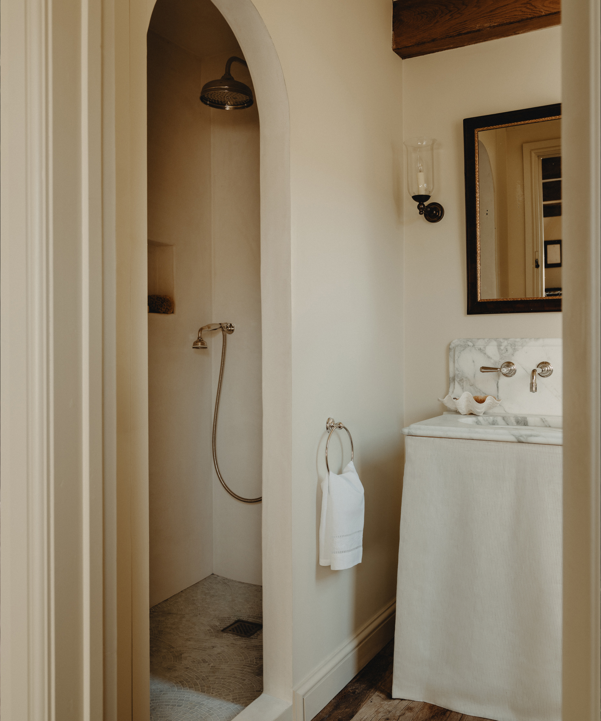 A small, elegant Berkeley-Hawkes bathroom with an arched walk-in shower, a wall-mounted showerhead, a towel ring with a white towel, a marble sink with a skirted base, and a mirror above the sink.
